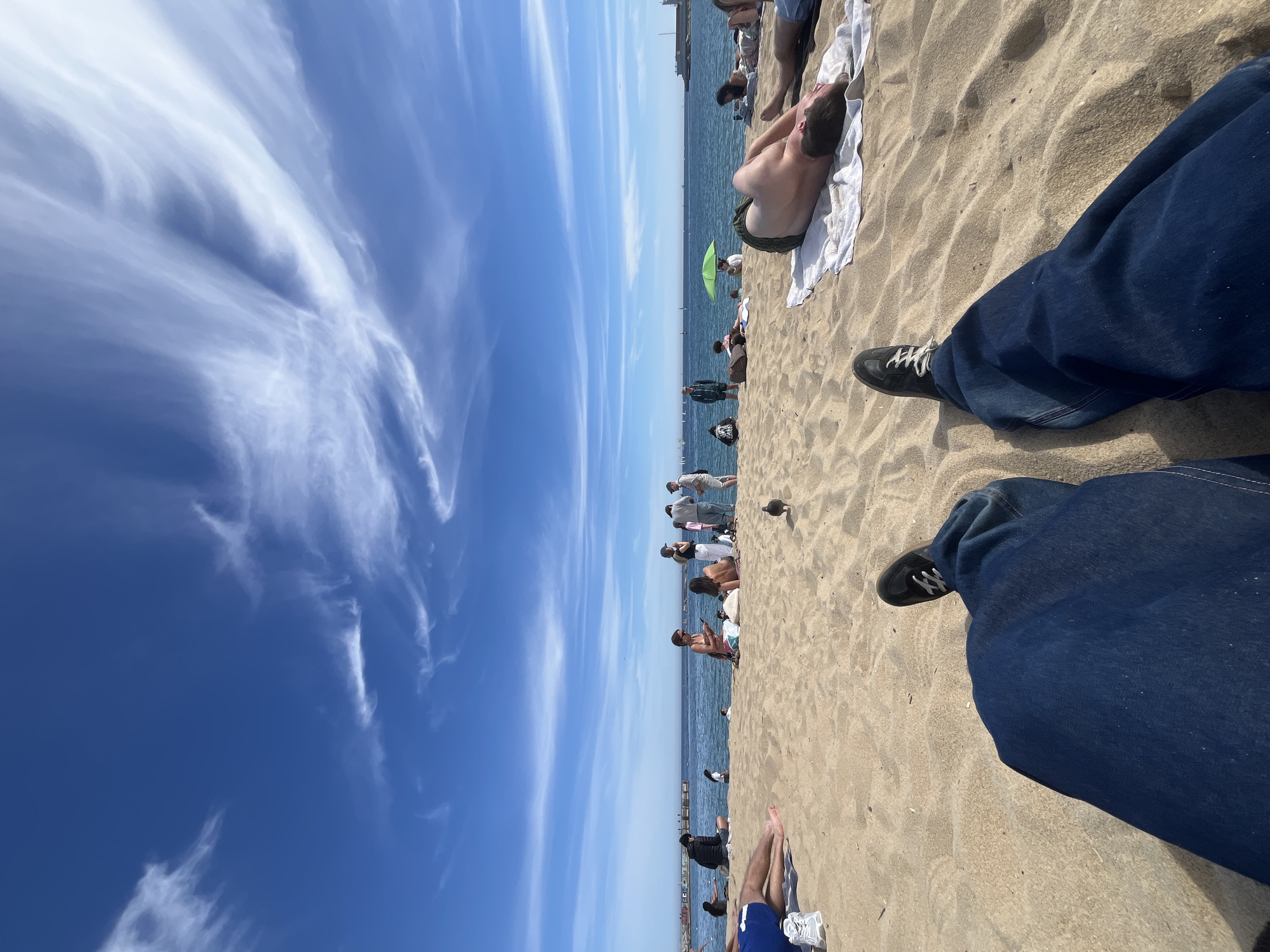 The scene is a beach in Barcelona, Spain, on a clear day with blue skies and wispy clouds. The perspective is from someone sitting on the sand, with their legs and feet in the foreground, clad in blue jeans and black sneakers with white laces.
The beach is moderately crowded with people sunbathing, walking, and relaxing. A man lies on a white towel in the sun. Further down the beach, there are groups of people, some standing, some sitting. The Mediterranean Sea is visible, separating the beach from the distant skyline of Barcelona, where a harbor can be seen.