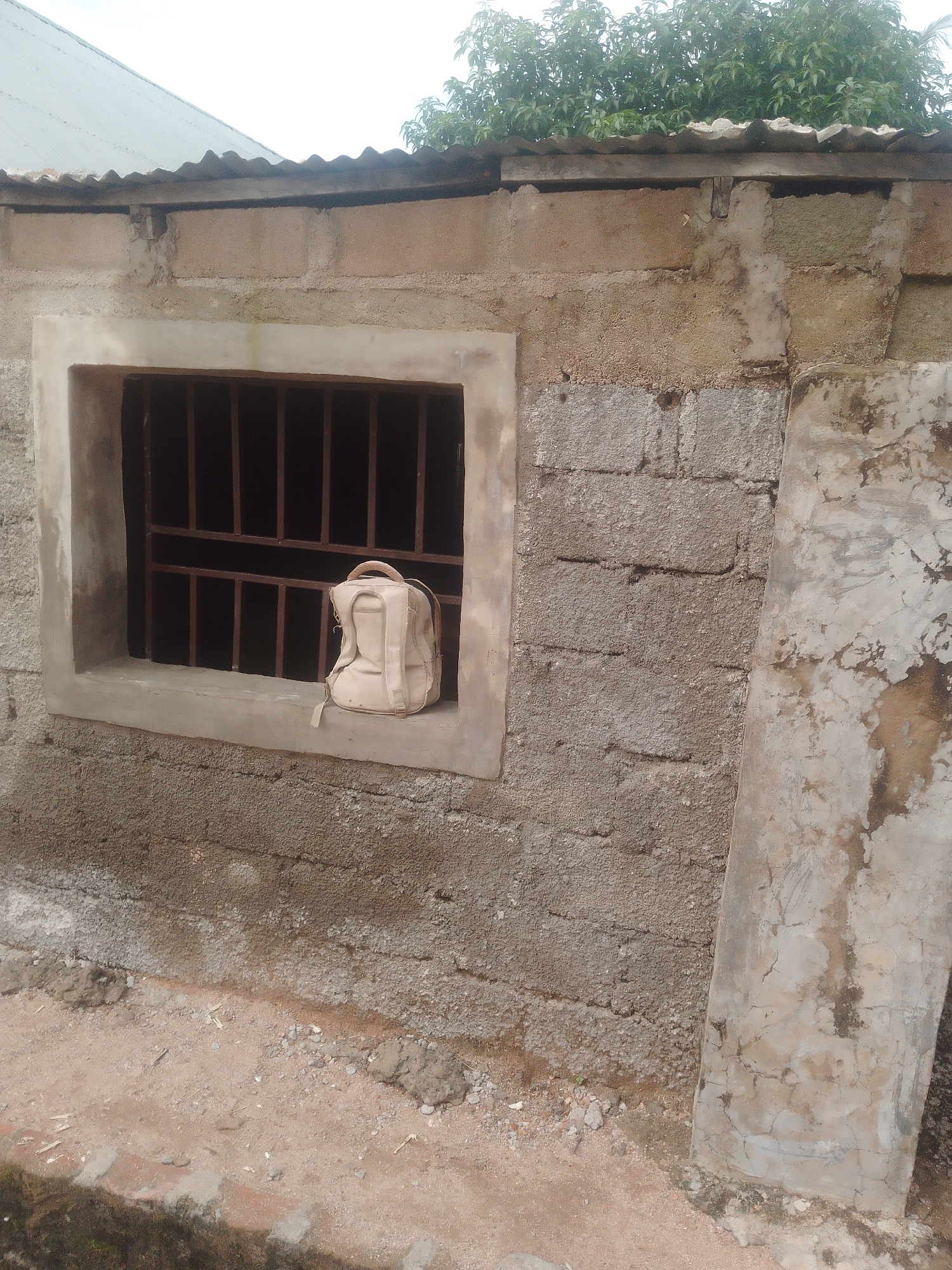 The image shows a close-up of a building's exterior in Jalingo, Nigeria. The building is constructed from concrete blocks and has a corrugated metal roof. A window opening is visible, reinforced with metal bars. A light-colored backpack rests on the windowsill inside the window. The ground in front of the building is covered in dirt and small stones. The overall impression is of a simple, utilitarian structure.