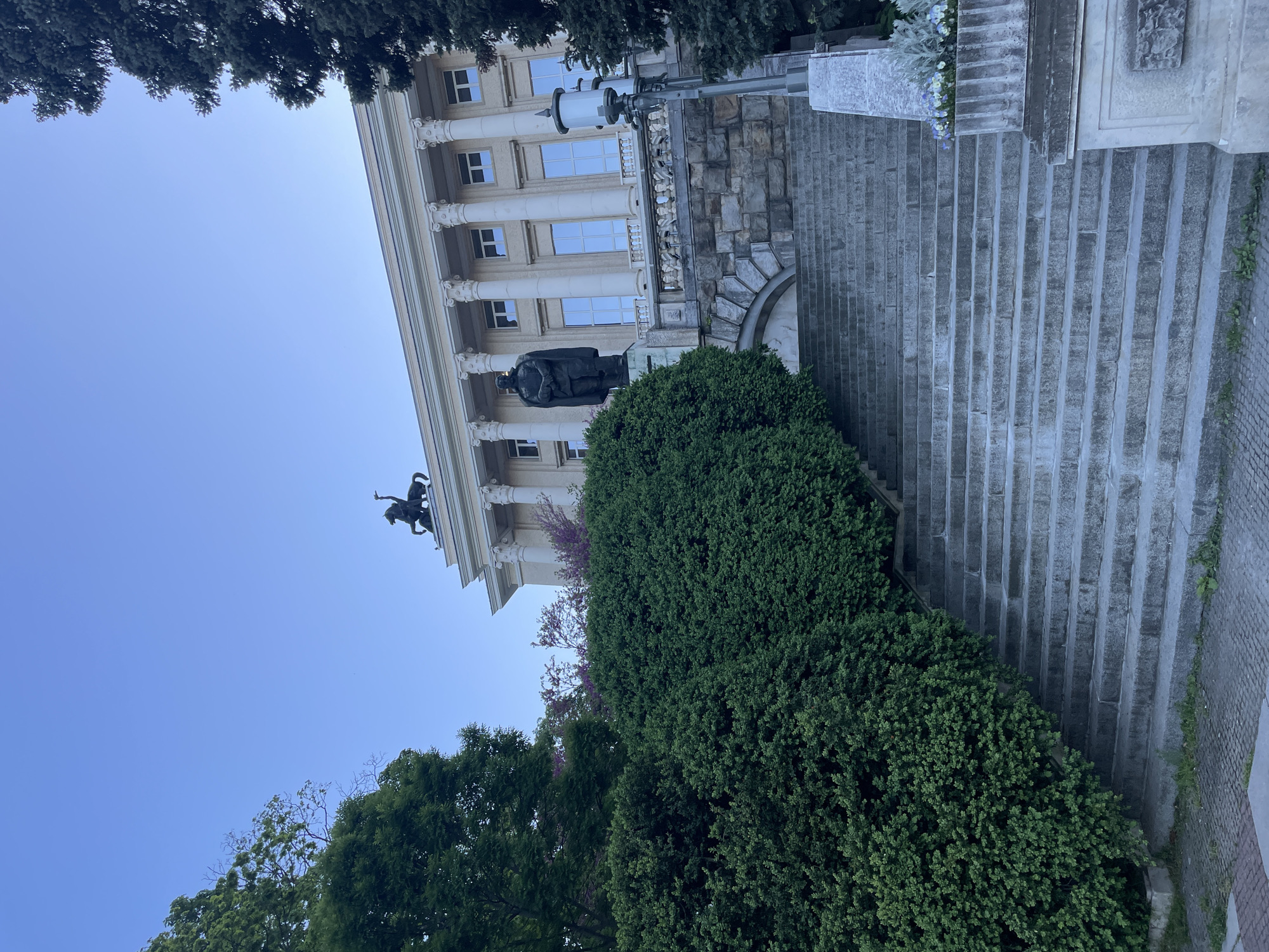 This photo shows the House of Humour and Satire in Gabrovo, Bulgaria. The building, featuring a neoclassical facade with columns, stands behind a statue and a green bush. A staircase is visible on the right side of the frame. The sky is light blue, and trees are visible in the foreground and at the top of the photo. The angle is a low-angle shot, tilted to the right.