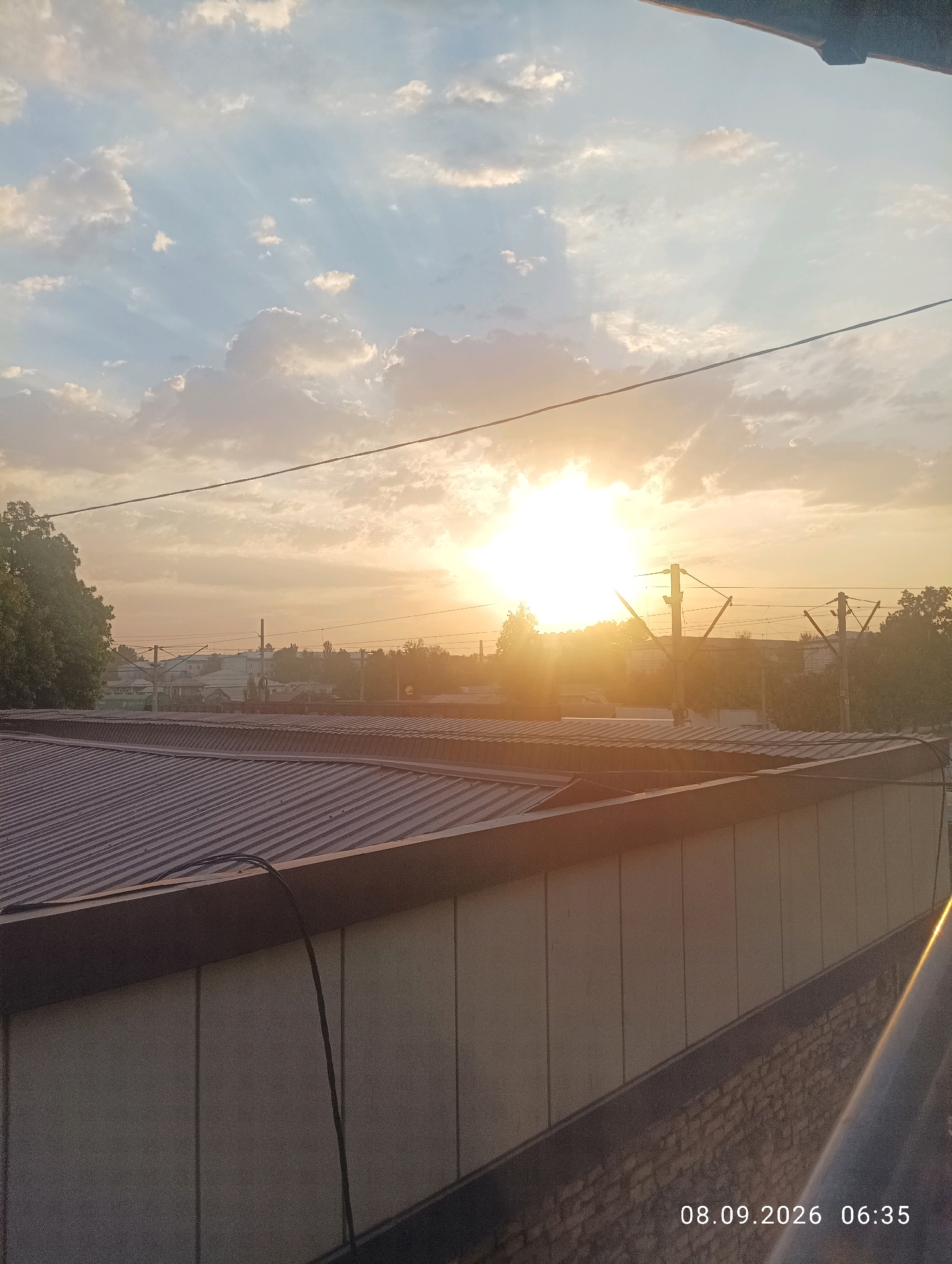 The image captures a sunset over a city, likely Yangiiul, Uzbekistan, as indicated by the date stamp "08.09.2026 06:35" which is in DD.MM.YYYY format. The sun is prominently visible, emitting bright rays through scattered clouds. In the foreground, a corrugated metal roof of a building is visible, with a beige facade below. A dark cable extends from the roof down the facade. In the midground, the silhouette of buildings, trees, and utility poles with overhead wires can be seen against the twilight sky. The overall scene depicts an urban environment during the golden hour. There are no people or animals visible in the image.