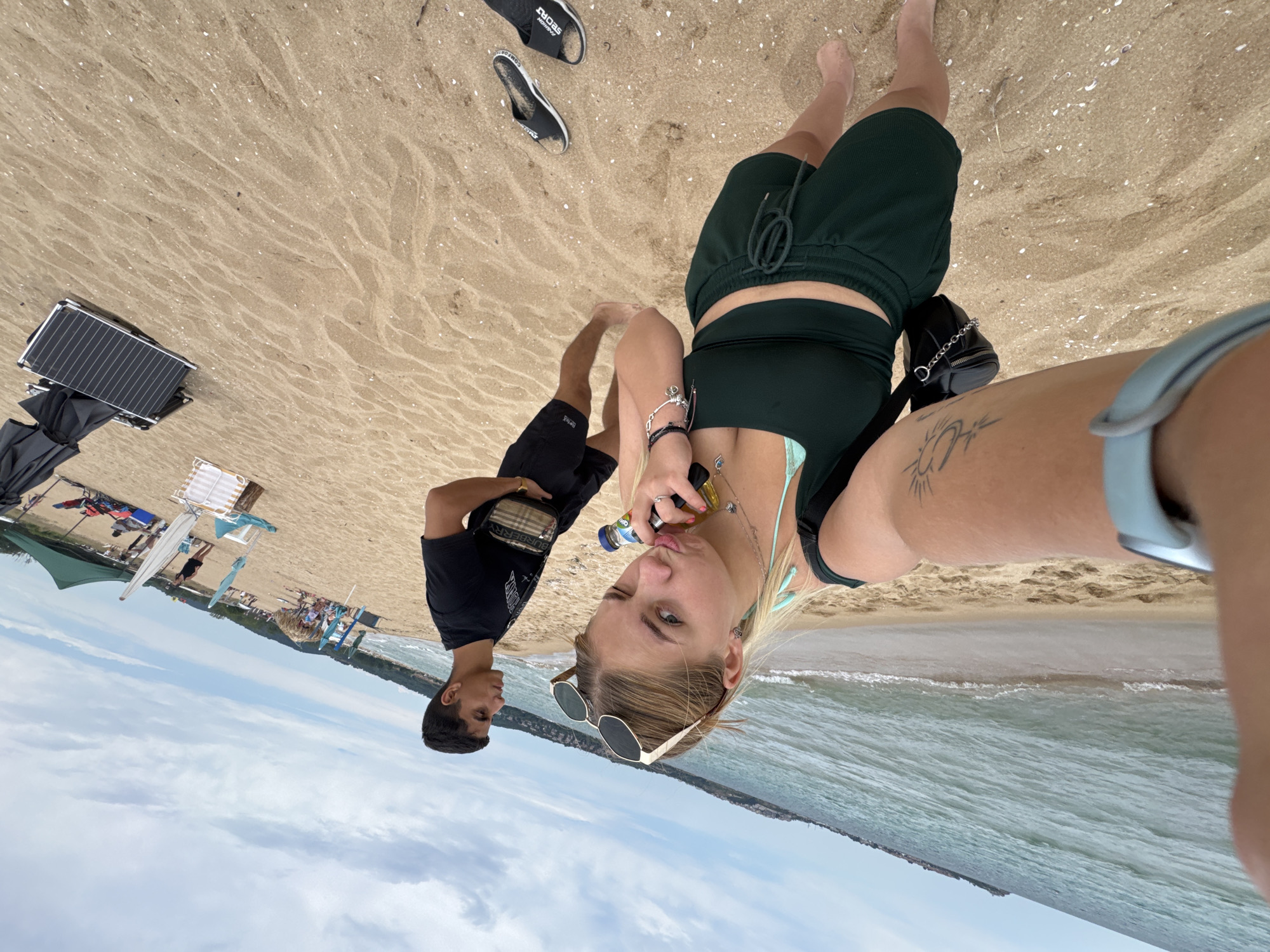The image depicts two young adults, a man and a woman, on a sandy beach with the ocean in the background. The man is wearing a black t-shirt and black shorts, with a crossbody bag. The woman is wearing a dark green halter top and matching shorts, with a blue bikini top underneath. She is holding a bottle and has sunglasses on her head. Both individuals appear to be posing for a selfie, with the camera angled to capture them and the surrounding beach environment.
The scene includes other beachgoers, beach chairs, and umbrellas further down the shore. The sand is light brown with scattered shells. The water is a light blue-green, with gentle waves lapping at the shore. The sky is cloudy, suggesting a potentially overcast or partly cloudy day. The overall atmosphere appears relaxed and casual, typical of a day at the beach.
A metal and black folding table or similar object is visible on the sand in the upper left portion of the image. There is also a black umbrella partially visible to the left of the folding table. Some of the beach chairs and umbrellas in the background have white and red or blue and white striped patterns.
Visible text includes "SPORTS" on the flip-flops near the top of the frame and "BURBERRY" on the man's crossbody bag.
The image is oriented with the horizon line appearing vertical, suggesting it was taken with the camera rotated. The time of day is not explicitly indicated but appears to be daytime, likely late morning or afternoon, given the lighting. The location is contextually identified as Sozopol, Bulgaria.