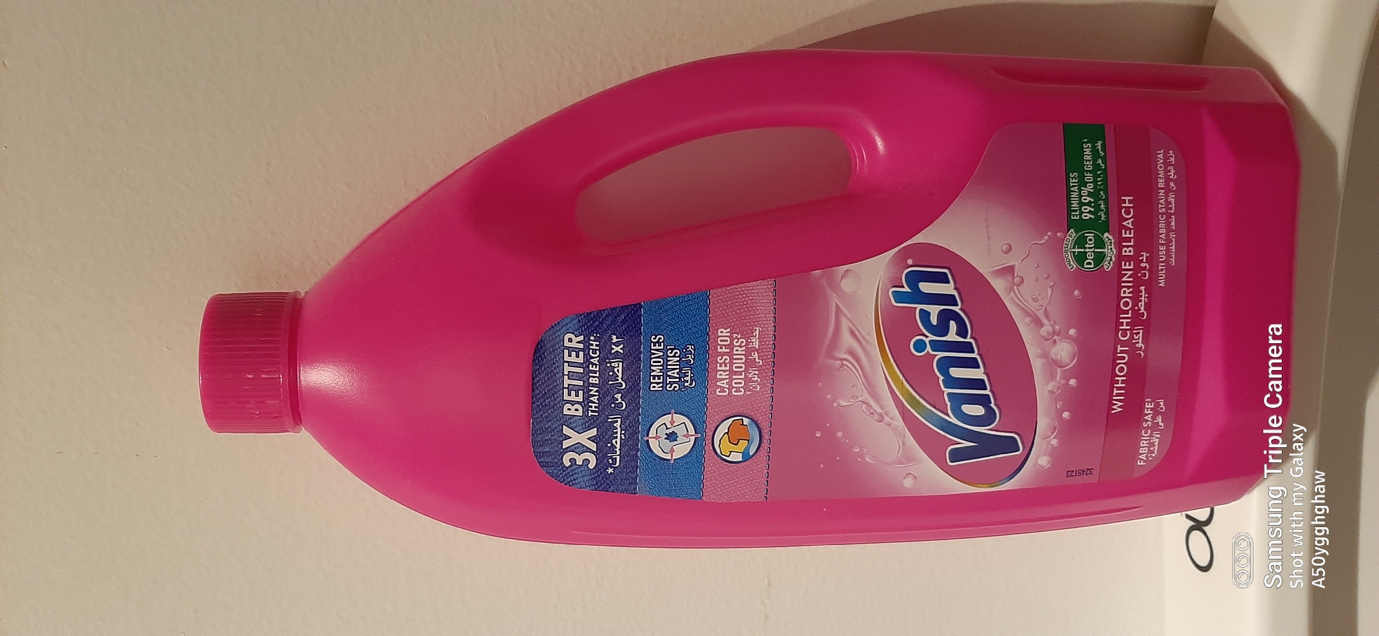 A bright pink plastic bottle of Vanish fabric stain remover is prominently displayed against a plain, light-colored wall. The bottle has a large handle on the right side and a pink screw-on cap. A label on the front of the bottle features the Vanish logo in large, stylized blue letters. The label also contains text in both English and Arabic, highlighting benefits such as "3X BETTER THAN BLEACH," "REMOVES STAINS," and "CARES FOR COLOURS." A smaller green circular logo from Dettol indicates that the product "ELIMINATES 99.9% OF GERMS." The bottom of the image is partially obscured by the text "Samsung Triple Camera Shot with my Galaxy A50ygghehaw." There is no visible indication of the city of Dubai, United Arab Emirates, or any specific urban environment. The scene appears to be indoors, with artificial lighting. No people or animals are present in the image.