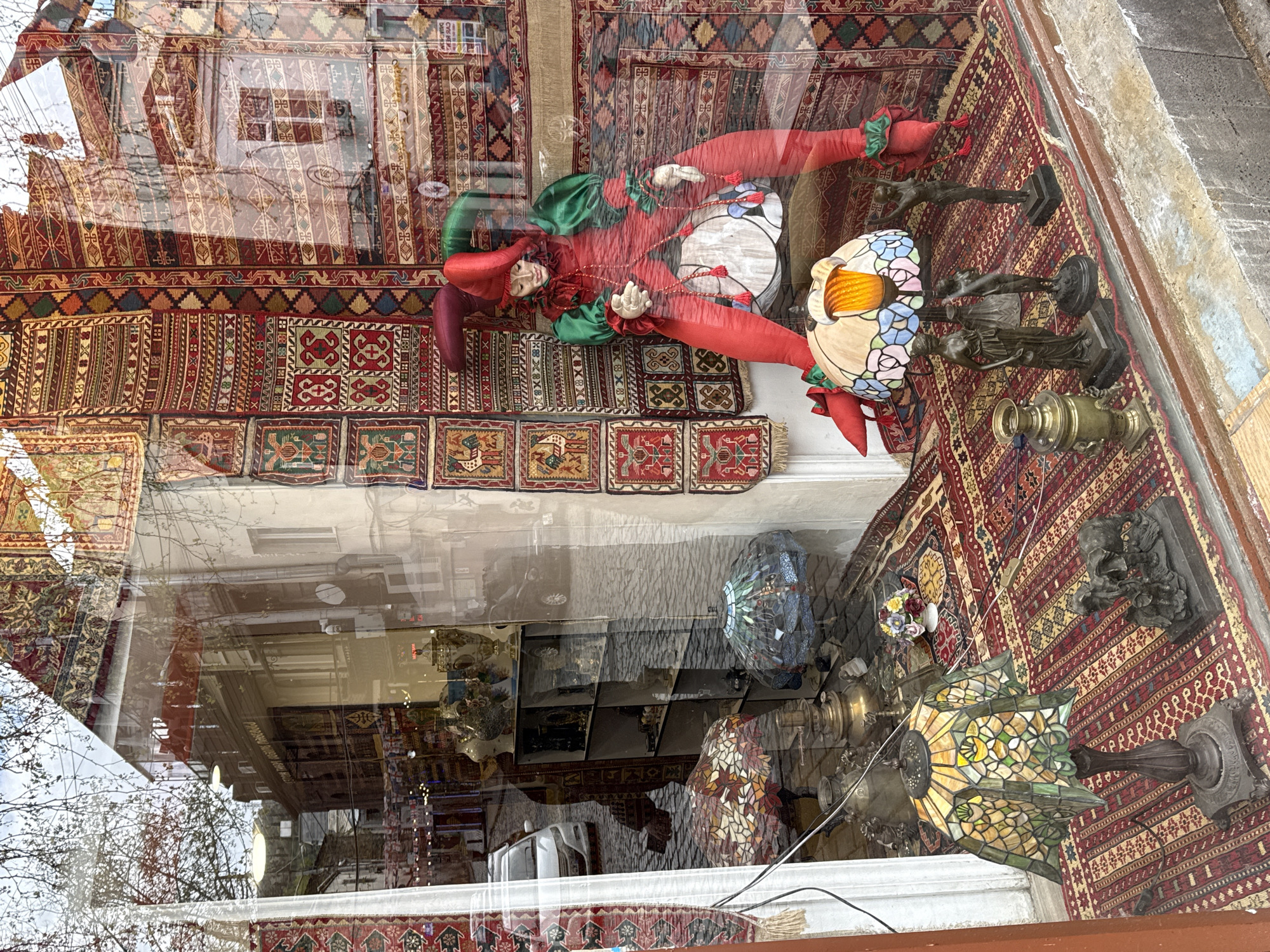 The image depicts the interior of a shop in Tbilisi, Georgia, viewed through a glass window that also reflects the street outside. The shop's display includes several items laid out on rugs: a red and green jester doll, several Tiffany-style lamps, and bronze sculptures. A samovar sits near one of the lamps. The rugs are patterned with reds and geometric designs. Reflections in the window show a building facade, possibly a street scene.