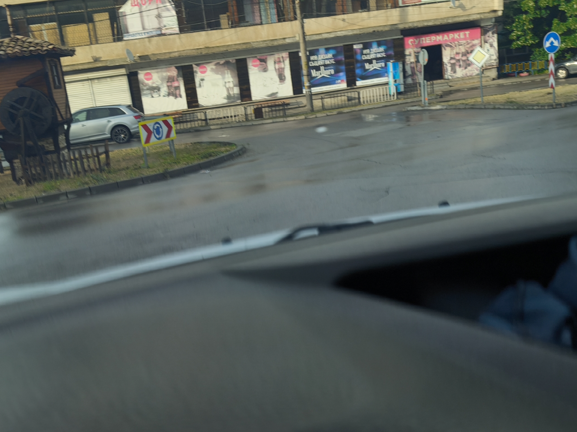 The image, captured from the perspective of a driver, depicts a wet street scene at an intersection or roundabout in what appears to be an urban or semi-urban environment. The road surface is visibly damp, suggesting recent rain, while the lighting indicates daytime, possibly morning or late afternoon.
In the foreground, the dashboard and a portion of the windshield and wiper of the vehicle from which the photograph was taken are visible.
The middle ground features a street with a grassy island on the left, adorned with a decorative wooden structure resembling a traditional water mill with a large wooden wheel. Behind this, a silver SUV is parked. A series of road signs are present: a yellow chevron sign with red arrows pointing right, directing traffic towards a blue circular sign with a white arrow forming a roundabout symbol, indicating a mandatory right turn. Further right, a yellow diamond-shaped sign with a white border denotes a priority road, and a blue circular sign with a right-turning white arrow is also seen.
In the background, a multi-story building lines the street. Its ground floor houses several storefronts. One prominent store entrance is marked by a pink banner with the Cyrillic word "СУПЕРМАРКЕТ" (Supermarket). Next to it, large billboards display text also in Cyrillic, reading "НОВ ДИЗАЙН СИЛЕН ВКУС" (New design strong taste) alongside the brand "Marlboro," indicating a tobacco advertisement. Other storefronts feature various promotional posters, some with indistinct images.
No individuals are clearly visible in the scene. The visible text and type of road signs suggest the location is in a Bulgarian-speaking country.