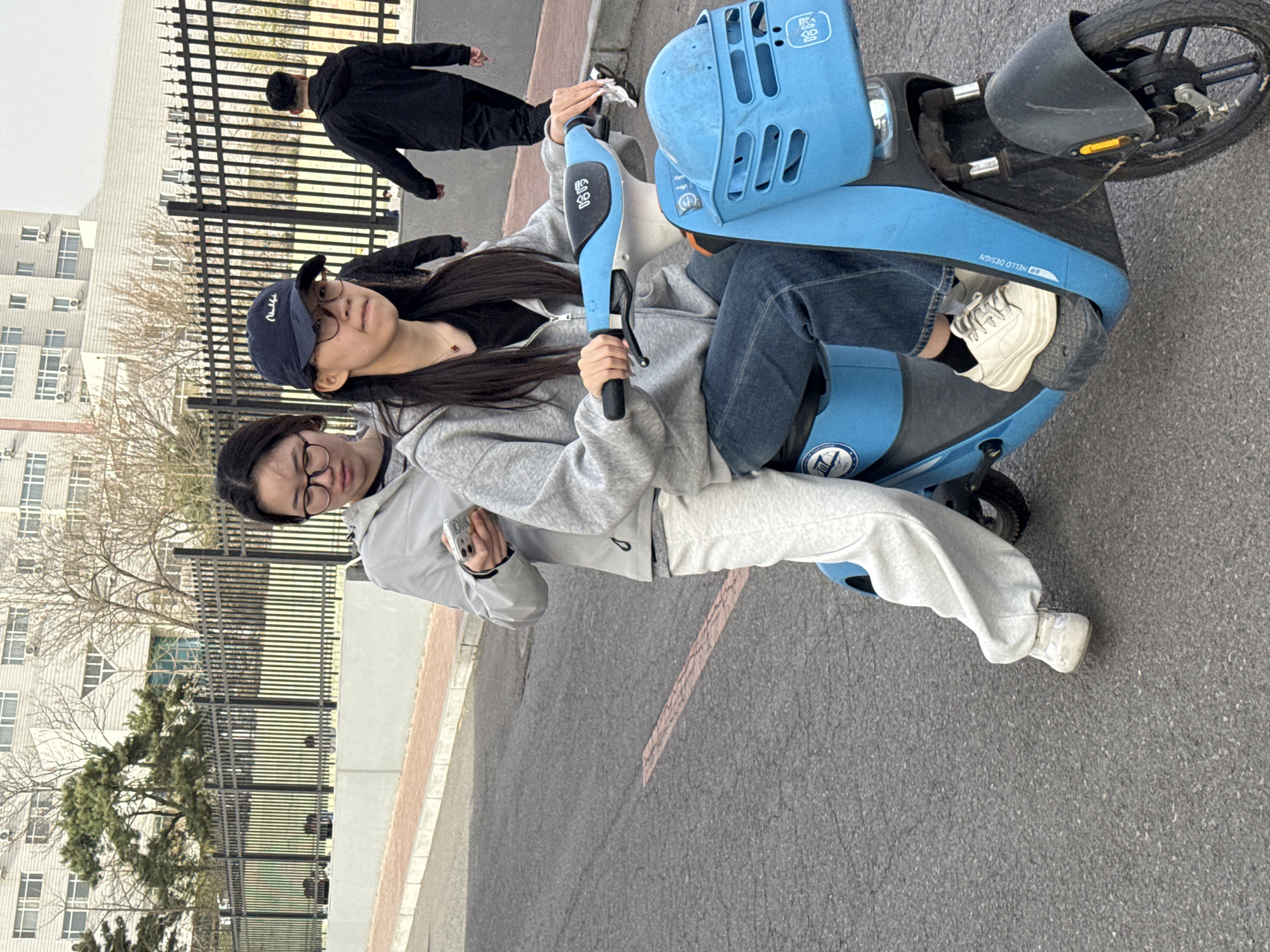 Two young women and a man are captured outdoors in a casual scene, likely in Zhongtun, China. The image is taken at a slight angle, with the subjects appearing rotated.
In the foreground, two young women are positioned around a bright blue electric scooter. One woman, wearing a dark baseball cap, glasses, a dark top, and blue jeans, is seated on the scooter, holding its handlebars. Her expression is neutral. Her left foot, in a white sneaker, rests on the scooter's footrest. The second woman, also wearing glasses, a light grey zip-up hoodie, and light grey sweatpants with white sneakers, stands beside the scooter, holding a mobile phone in her left hand. She has long dark hair and a neutral expression.
The blue electric scooter features a front basket with Chinese characters, and text on its side that includes "HELLO DESIGN" and possibly a model number like "868i." Additional Chinese characters are visible on the main body of the scooter, likely branding for a shared bike service.
In the background, behind a dark metal fence, a man in a black hoodie and black pants walks away from the camera. Further back, there are light-colored buildings with numerous windows and visible air conditioning units, suggesting an urban or suburban setting. Several trees, some bare and some with green foliage, are scattered behind the fence. The ground is an asphalt road with a concrete sidewalk featuring reddish-brown tiles.
The time of day appears to be daytime, with soft, diffused lighting, suggesting an overcast sky or a time of day without direct harsh sunlight. The overall atmosphere is relaxed and candid.