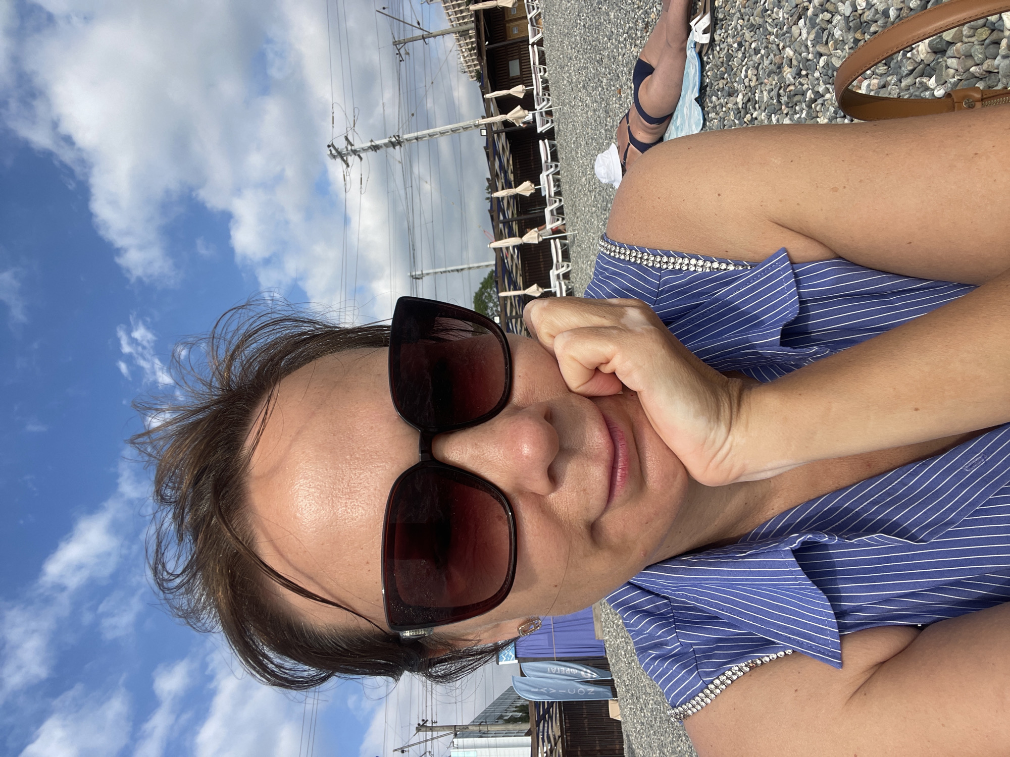 A woman with brown hair and sunglasses is taking a selfie on a pebble beach in Sochi, Russia. She is wearing a blue and white striped top adorned with rhinestones on the shoulders. Her right hand is supporting her chin as she looks at the camera. In the background, beach umbrellas and lounge chairs are set up, and another person is lying on a towel. Power lines and some buildings are visible further in the distance, suggesting an urban coastal setting. The sky is blue with scattered white clouds.