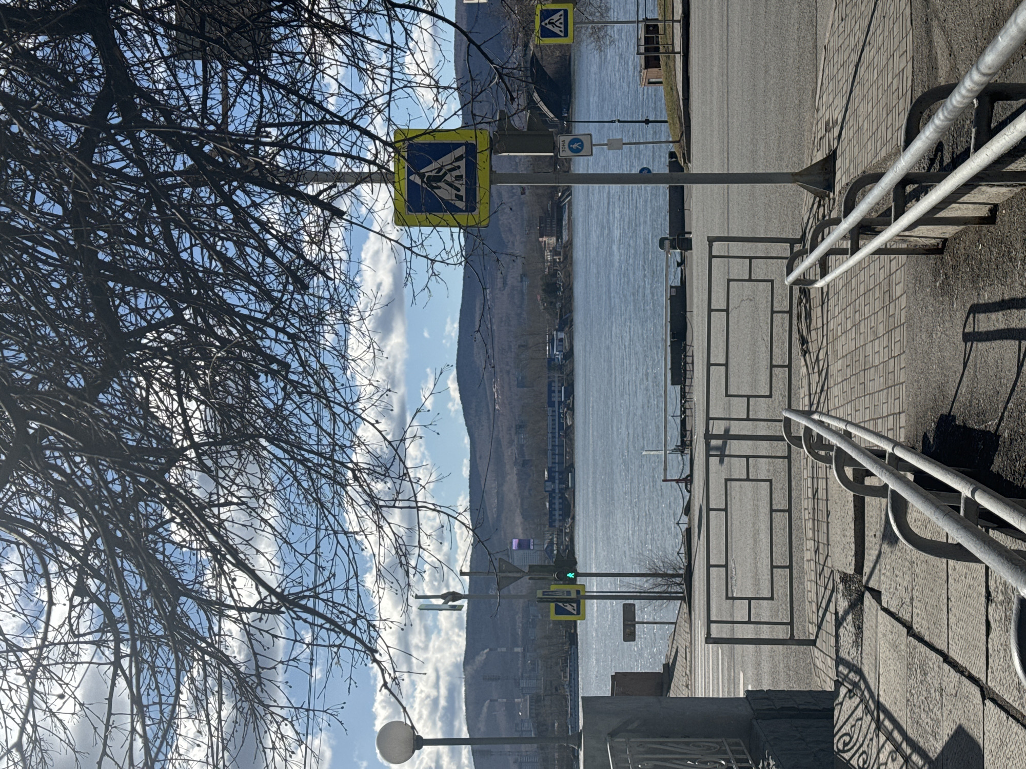 The image shows a waterfront scene in Krasnoyarsk, Russia, near the Yenisey River.
**Environment:** The scene is set alongside the Yenisey River, with views of the opposite bank, including buildings and mountainous terrain in the background. The sky is blue with scattered clouds. There's a tree with bare branches in the left foreground.
**Objects:** Prominent objects include:
* Two pedestrian crossing signs (yellow and blue), one near the foreground and another further along the walkway.
* A green traffic light.
* A sidewalk with metal railing.
* A metal fence along the walkway.
* A lamppost.
**People/Actions/Interactions:** There is no people present in the image.
**Scene details:** The image appears to have been taken from a slightly elevated position. The pedestrian crossing signs and the traffic light are indicators of a road crossing point. The metal railing and fence suggest a barrier or safety measure along the walkway.