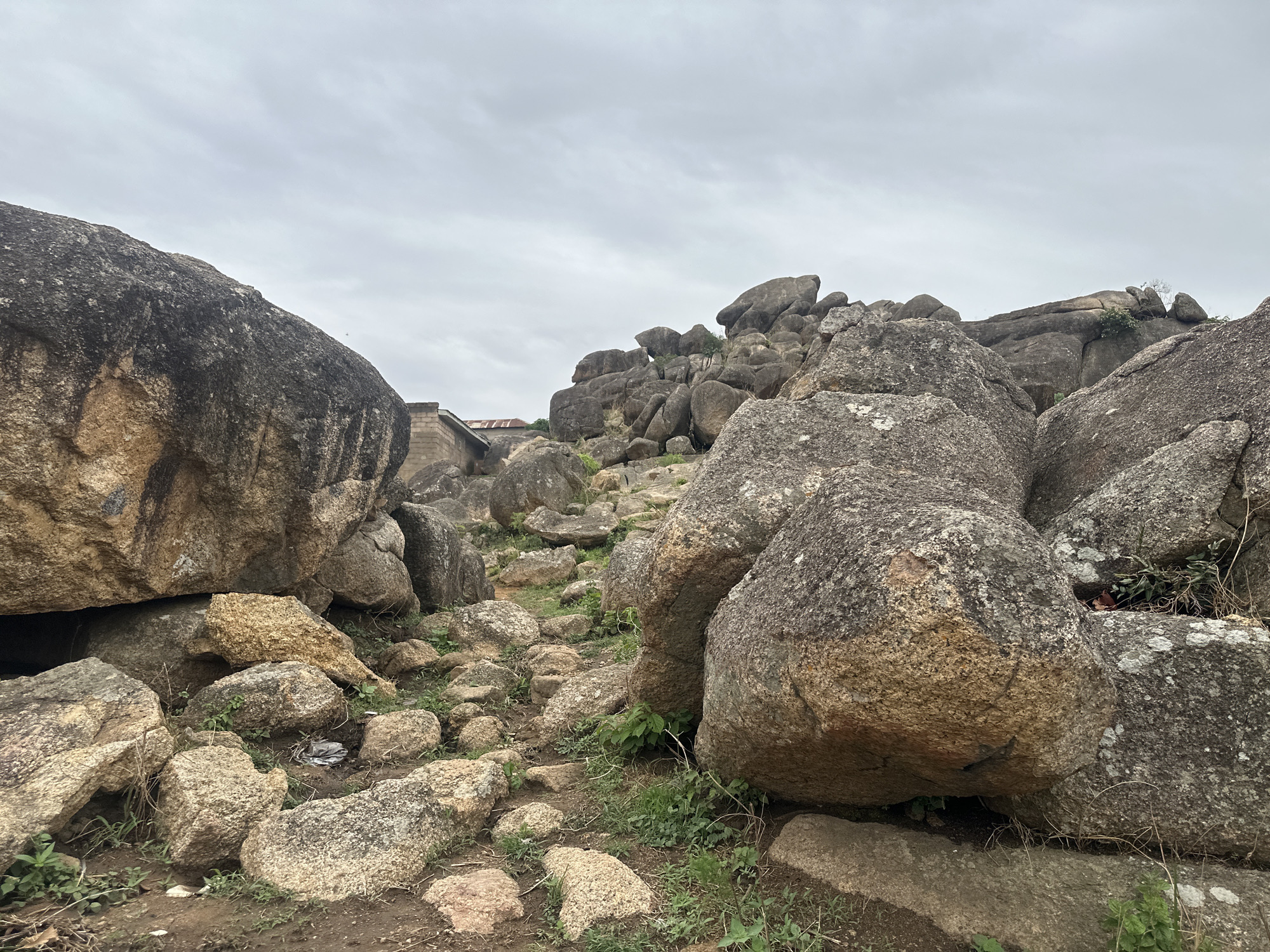 The image depicts a rocky landscape in Jos, Nigeria. Large granite boulders dominate the scene, interspersed with smaller stones and sparse vegetation. A path winds through the rocks, leading towards a small building with a corrugated metal roof in the distance. The sky is overcast, suggesting a cloudy day. No people or animals are visible.