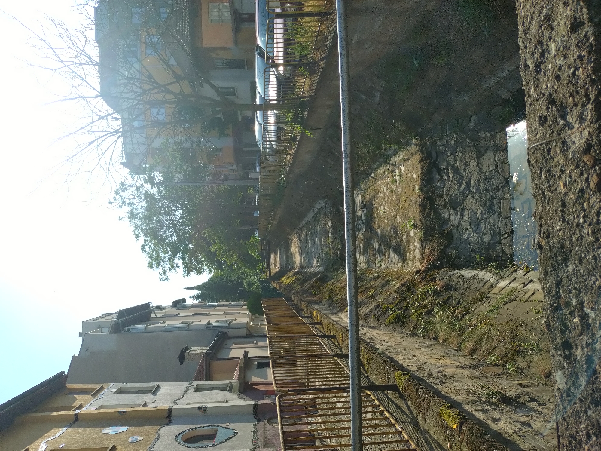 This image depicts an urban canal scene in Stara Zagora, Bulgaria, viewed from a slightly elevated position. The canal, appearing largely dry or with very low water, runs from the foreground towards the background, flanked by stone and concrete retaining walls that show signs of moss and plant growth. A small pool of water is visible at the lowest point of the canal bed, reflecting bright sunlight.
On the left side of the canal, a rusty golden-brown metal fence runs along a raised concrete pathway. Behind this fence, several multi-story buildings are visible. The closest building features a distinctive yellow-brown facade adorned with circular and hexagonal decorative mosaic elements. Further down the street, additional buildings with plainer grey and beige facades are present.
On the right side of the canal, another identical rusty golden-brown metal fence borders a parking area or street. Behind this fence, a line of various parked cars (including sedans and hatchbacks) is visible, their surfaces reflecting the bright sunlight. Beyond the cars and fences, multi-story buildings with yellow and grey facades are present, partially obscured by a mix of leafy and bare-branched trees.
The sky in the upper portion of the image is bright and appears overexposed in areas, indicating strong direct sunlight. Shadows are cast towards the left side of the frame. Two small pedestrian bridges or crossings are visible spanning the canal further in the background. The immediate foreground shows a rough, textured concrete surface from which the photo was taken, with a horizontal metal pipe crossing the mid-ground. No people are visible in the scene.