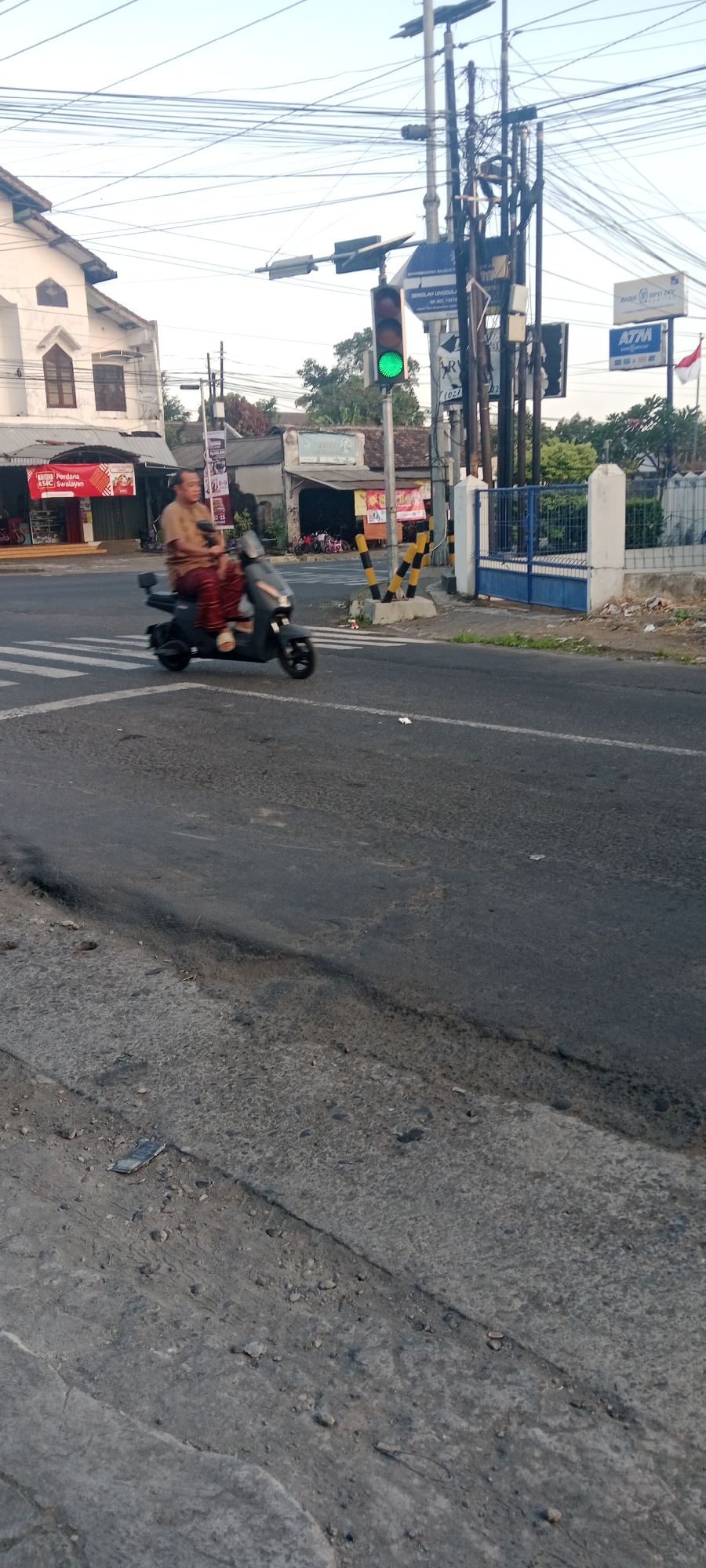 A man is riding a dark-colored scooter at an intersection in Maguwoharjo, Indonesia. The traffic light is green. The man is wearing a light brown long-sleeved shirt and patterned pants. He is looking forward. The scooter has a small windshield. The intersection has a pedestrian crossing marked on the asphalt. In the background, there is a white building with a sloped roof on the left, and various buildings and structures on the right, including utility poles with many wires, a blue metal gate, and a sign that says "BANK BPD DIY". Trees are visible behind the buildings. The sky is clear and light. The road surface is asphalt, with some visible cracks and debris.