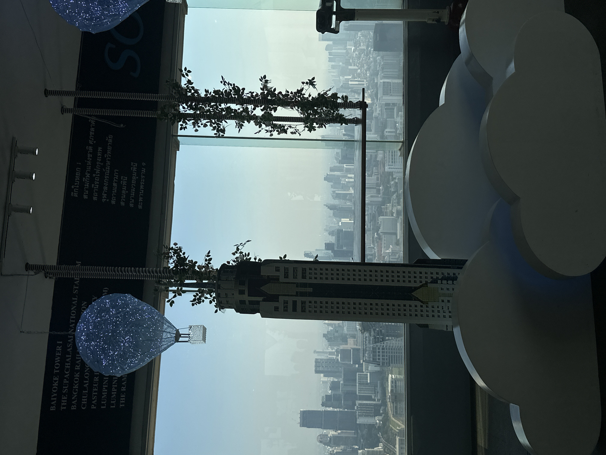 The scene is taken from the interior of the Baiyoke Tower I in Bangkok, Thailand, as identified by signage visible in the image.
The view faces outward through a glass window overlooking the cityscape. In the foreground, there are interior decorative elements, including a small model or representation of a building adorned with artificial plants. Beside this model, a decorative cloud-shaped sculpture is visible. To the left, a sign includes the name of the location. Above the sign, a decorative illuminated hot air balloon-shaped light fixture hangs.
Outside the window, a dense urban landscape is apparent, showing numerous buildings varying in size and height under a hazy or smoggy sky. The overall atmosphere suggests a busy and densely populated area.