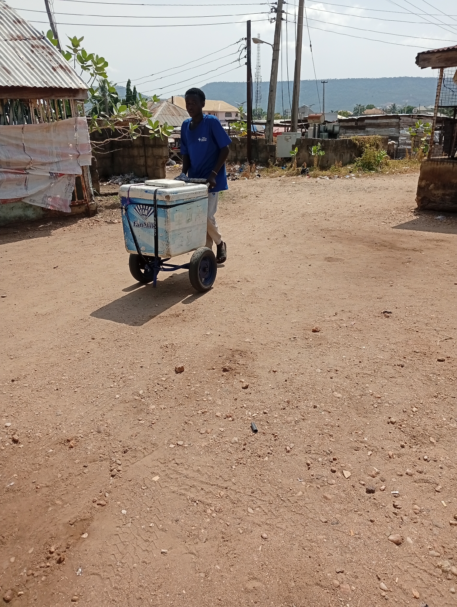 A young man pulls a wheeled cart bearing a white, insulated cooler through a dirt street in Adankolo, Nigeria. The cooler is branded with "Fan Milk" logos. The man wears a blue t-shirt, light-colored pants, and dark shoes. The ground is reddish-brown dirt mixed with stones and tire tracks. A corrugated metal structure is on the left side of the frame. Debris and structures are visible in the background. Power lines and distant hills are visible. The sky is overcast.