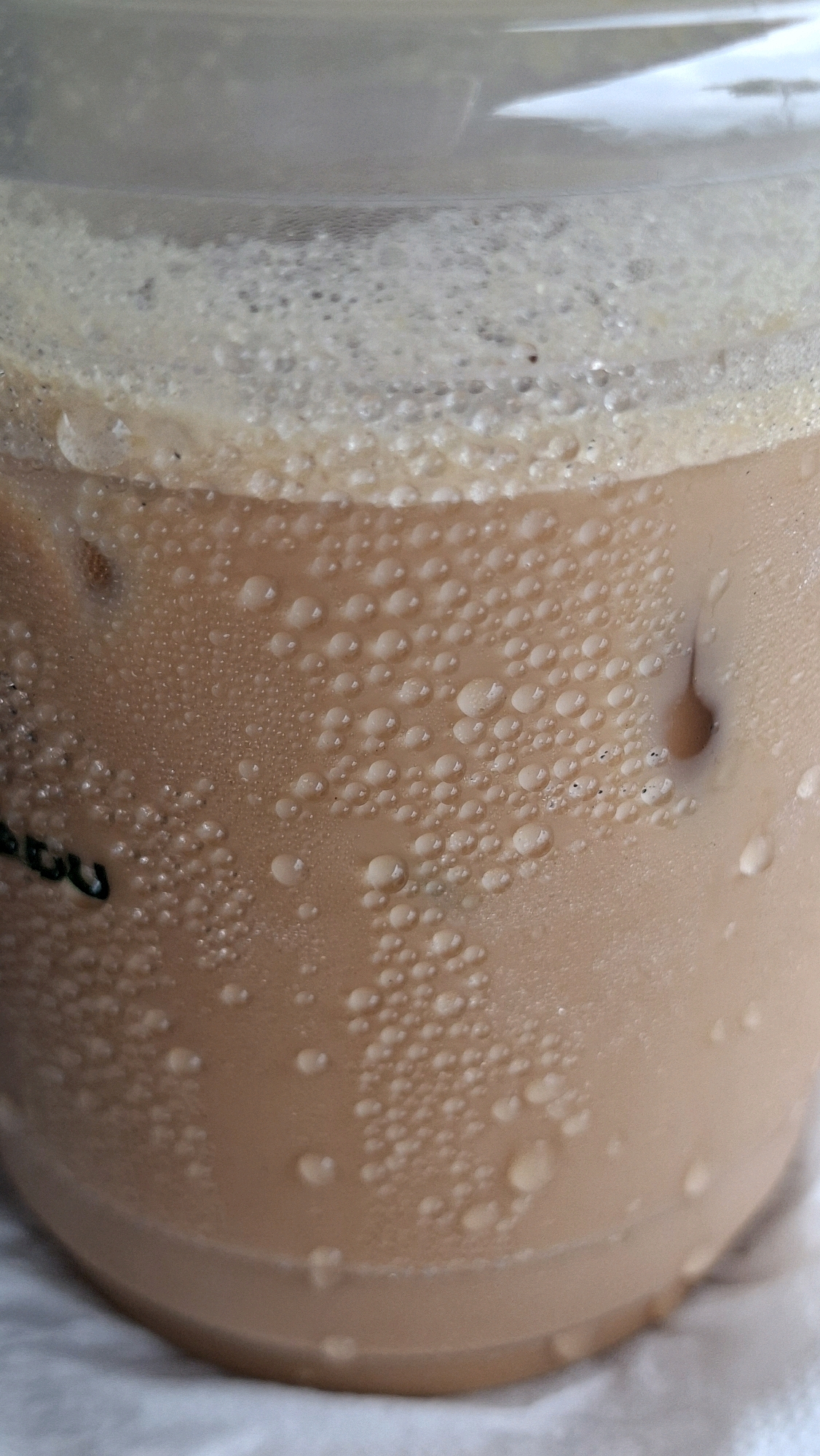 The image shows a close-up of a clear plastic cup filled with a light brown liquid, likely iced coffee or tea. The exterior of the cup is covered in condensation droplets of various sizes, indicating a cold beverage. Some ice cubes are visible through the translucent liquid. At the top of the beverage, there is a layer of foam. There are no people or any discernible landmarks to indicate the location within Keudebayu, Indonesia. The focus is entirely on the condensation and the beverage itself.
