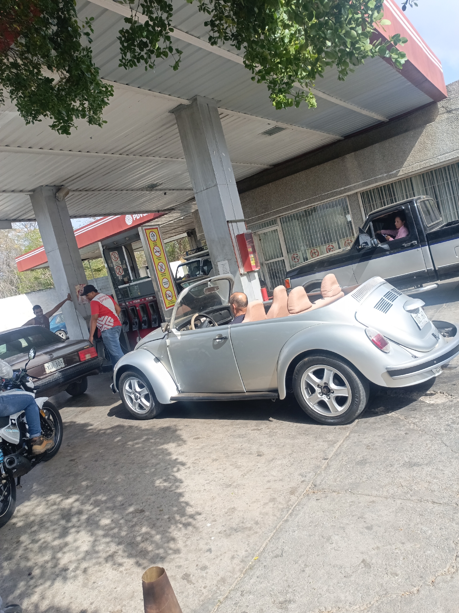 The scene depicts a gas station in La Tagua, Venezuela. A silver convertible Volkswagen Beetle is parked at a pump with a man sitting in the driver's seat. Another vehicle, a dark-colored sedan with license plate "XDR-103," is also at the pump, being attended to by a person in a red and white shirt. A man is standing to the left of the sedan. A motorcycle is visible in the foreground. A black pickup truck with a woman in the driver's seat is parked nearby. The gas station has a canopy supported by concrete pillars. A "PDVSA" sign is visible on the gas pump. The ground is concrete and shows signs of wear. There are trees visible in the background and on the left of the frame.