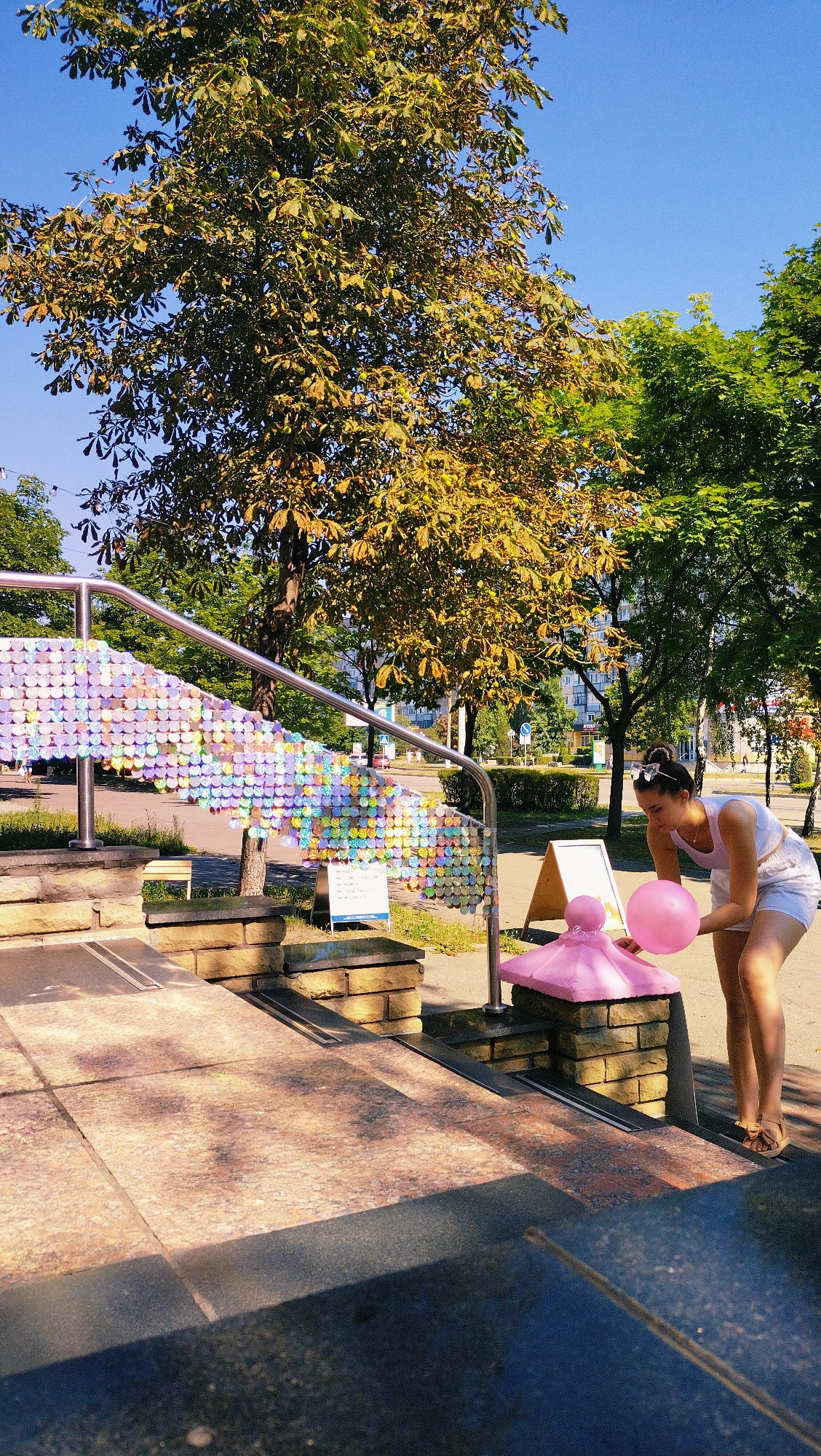 The image shows a woman in a white tank top and shorts holding a pink balloon, standing on the steps of an outdoor staircase in the city of Horishni Plavni, Ukraine. The staircase is decorated with a shimmering, colorful sequin-like material on its railing, creating a wave-like pattern. The woman is looking down at a pink, pedestal-like object near the bottom of the stairs. Behind the stairs, a large tree with yellowing leaves dominates the background, with a clear blue sky above. In the distance, buildings and street signs are visible, indicating an urban environment. The lighting suggests it is a sunny day.