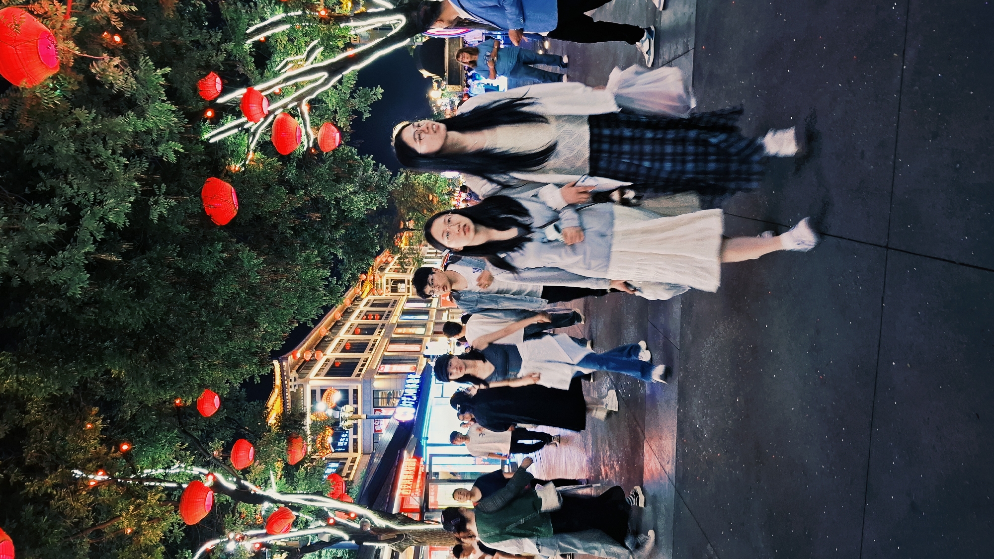 The image captures a nighttime scene in Yanta District, China, with a focus on an urban street adorned with illuminated trees and red lanterns. A group of people, predominantly young adults, are walking along the street, their forms blurred, suggesting motion. Two women stand out, appearing more still compared to the others, both dressed in light-colored tops and skirts, and engaged in conversation.
In the background, buildings with traditional Chinese architectural elements are visible, illuminated with warm lights, which accentuates their decorative features. The lanterns hanging from the trees provide a festive and cultural ambience. The pavement appears clean, reflecting some of the ambient light. The composition is slightly tilted.