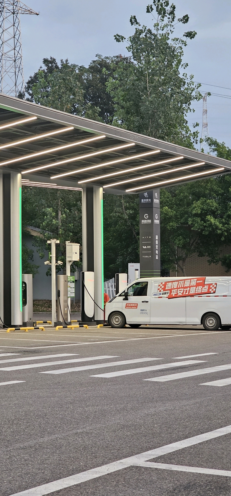 In Longqiao, China, a white van is parked at a charging station under a covered structure with LED lighting. The van has red and white lettering on its side. The charging station features multiple charging stalls, some with green lights illuminated. The ground in front of the station is paved with asphalt and marked with white pedestrian crossing lines. Trees and power transmission towers are visible in the background. The sky is overcast.