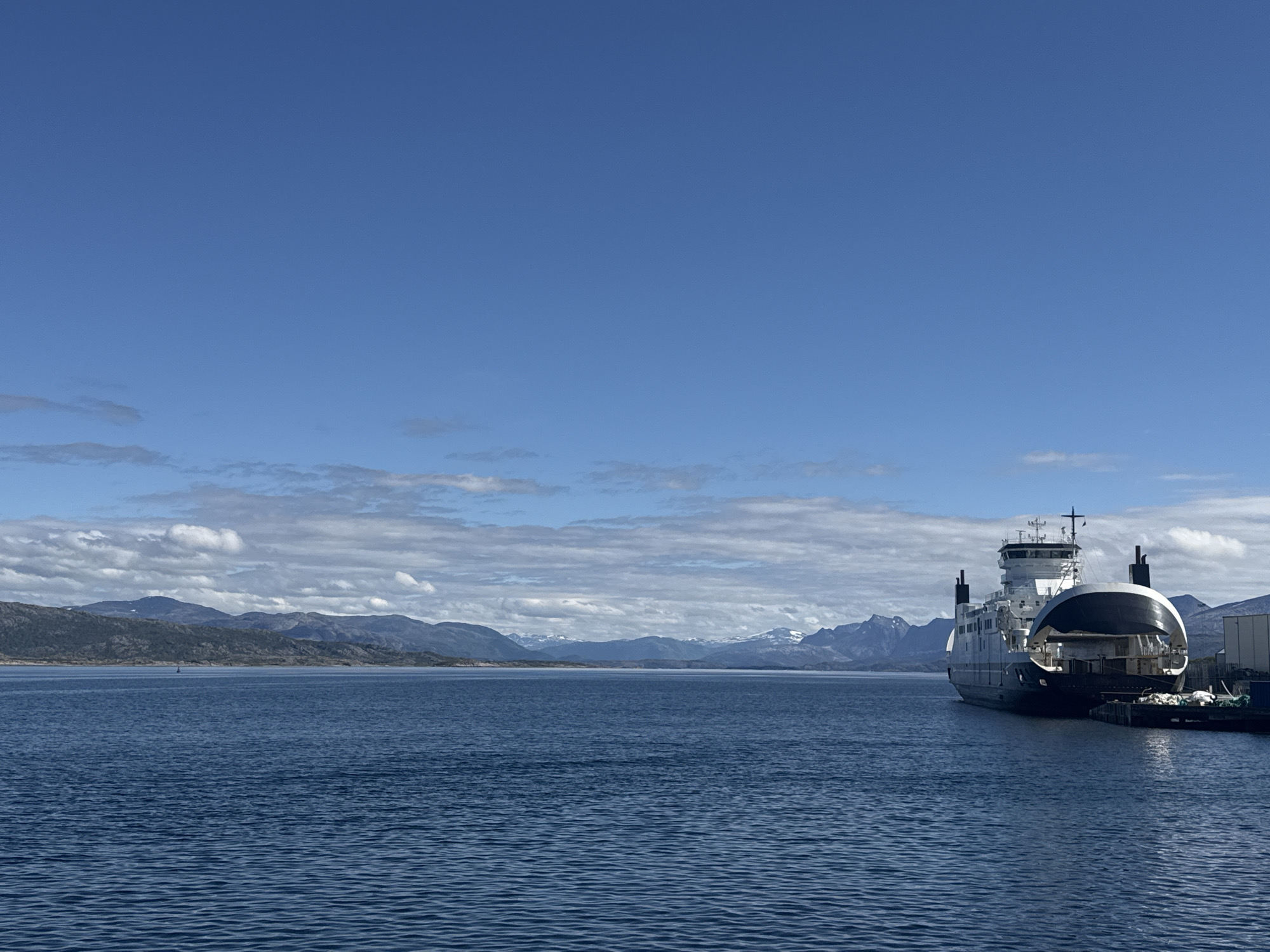 The image shows a ferry docked at a pier in Lødingen, Norway. The ferry, which is primarily white and black, is positioned on the right side of the frame, facing towards the left. It has a prominent upper structure with windows and what appears to be a bridge. The ferry's open bow ramp suggests it is ready for loading or unloading. The water in front of the ferry is a deep blue and appears calm with small ripples.
In the background, across the water, a range of mountains is visible. Some of the peaks are snow-capped, indicating a mountainous terrain likely characteristic of the Lofoten Islands region, where Lødingen is situated. The sky is a clear, vibrant blue with scattered white clouds, particularly concentrated near the horizon above the mountains. The overall lighting suggests it is daytime, with the sun casting gentle light on the scene.
The pier area to the right of the ferry shows some industrial elements, including what looks like a building or storage facility, along with some equipment and ropes. There are no people visible in the image, nor are there any other vessels in the immediate vicinity besides the docked ferry. The scene conveys a sense of a functional port in a scenic, coastal environment.