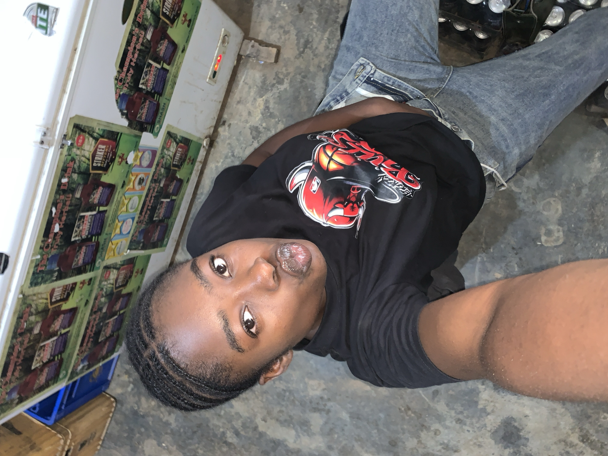 A young Black person with braided hair is lying on a concrete floor next to a white chest freezer. The freezer has "STRIKER" herbal bitters advertisements on its side. The person is wearing a black t-shirt with a "Chicago Bulls" logo and blue jeans. A case of drinks can be seen in the background next to the jeans. The photo may have been taken at the New Stadium in Nigeria.