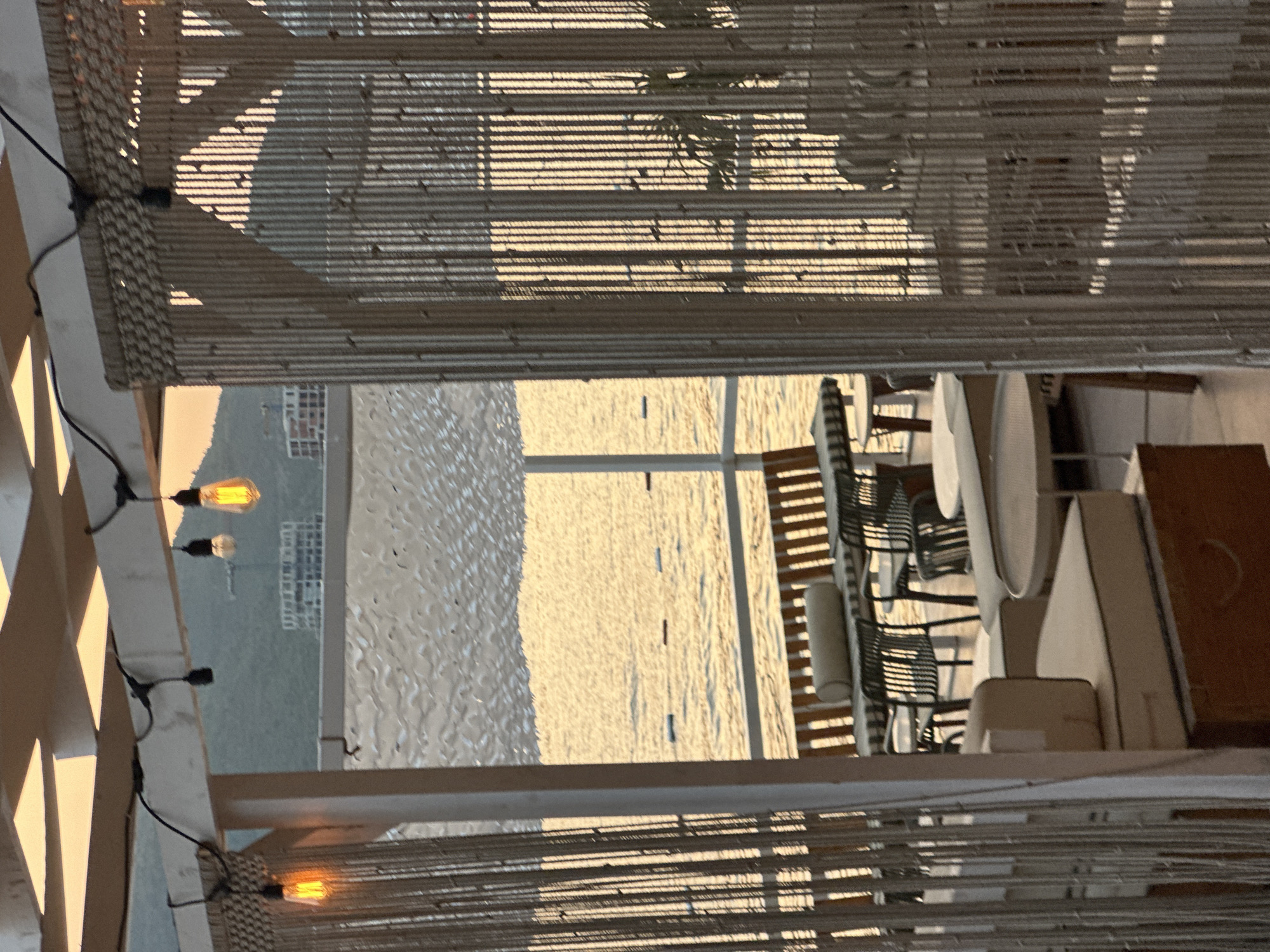 The image depicts an outdoor seating area with a view of the sea during sunset. In the foreground, a beaded curtain hangs, obscuring part of the scene. Beyond the curtain, a covered patio features seating arrangements, including cushioned sofas, tables, and chairs. The sea is visible in the middle ground, with gentle waves and a few buoys marking an area. In the background, a hillside with buildings is silhouetted against the orange-hued sky, indicative of a coastal city. The overall impression is one of a tranquil, seaside establishment in Budva, Montenegro, likely a restaurant or cafe, as evidenced by the outdoor dining setup.