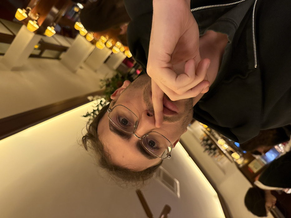 A person wearing glasses and a black hoodie looks into the camera, their finger touching their lip. The setting is a dimly lit restaurant, evident by the soft lighting and table settings in the background. Other patrons are partially visible in the blurred background. The image orientation is rotated, with the table appearing vertical.