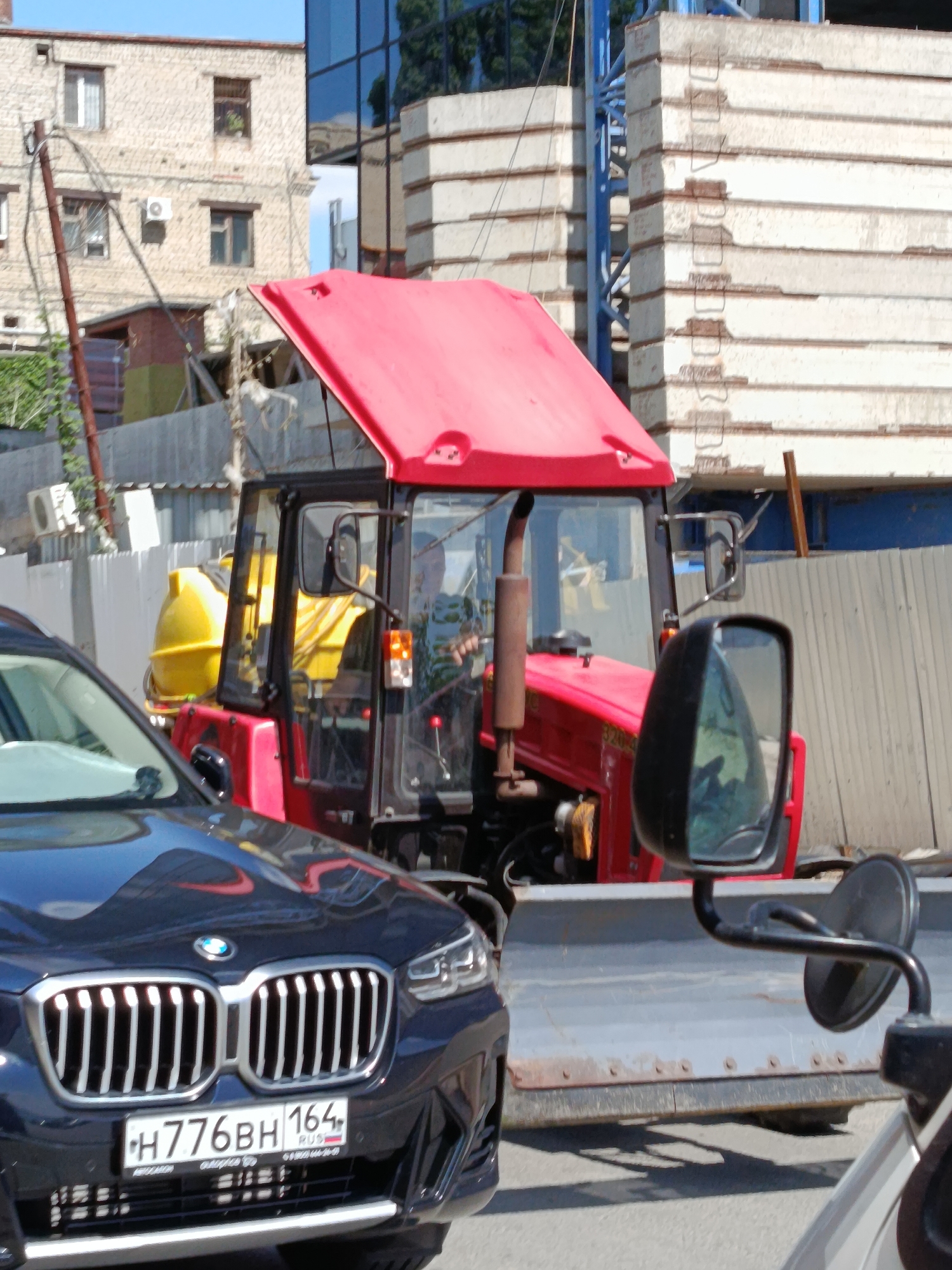 A dark blue BMW SUV is parked in front of a red tractor with a snowplow attachment. The tractor has a yellow cylindrical tank on its back. The scene appears to be in an urban environment, with buildings visible in the background. The buildings are made of brick and glass, and there is a construction crane in the distance. The license plate of the BMW is visible and reads "H776BH 164 RUS". The image was taken in the city of Saratov, Russia.