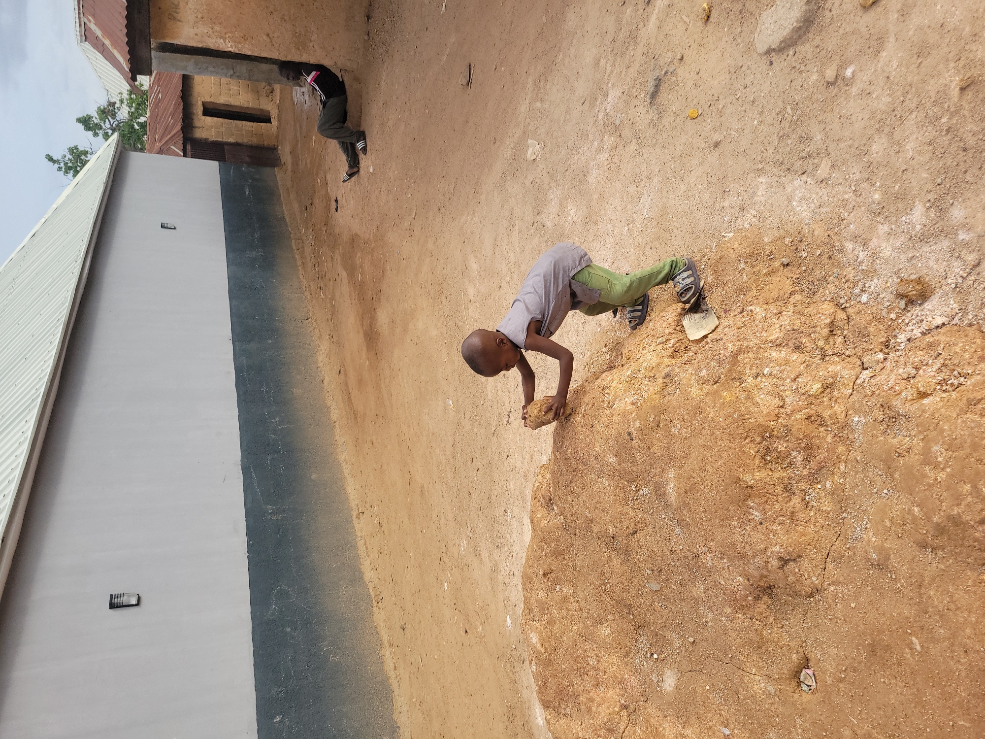The image depicts an outdoor scene in Jos, Nigeria. Two children are present. One child is squatting down and is holding a rock. The other child walks toward the left corner of the image. There is an adjacent wall and a dirt/rock terrain.