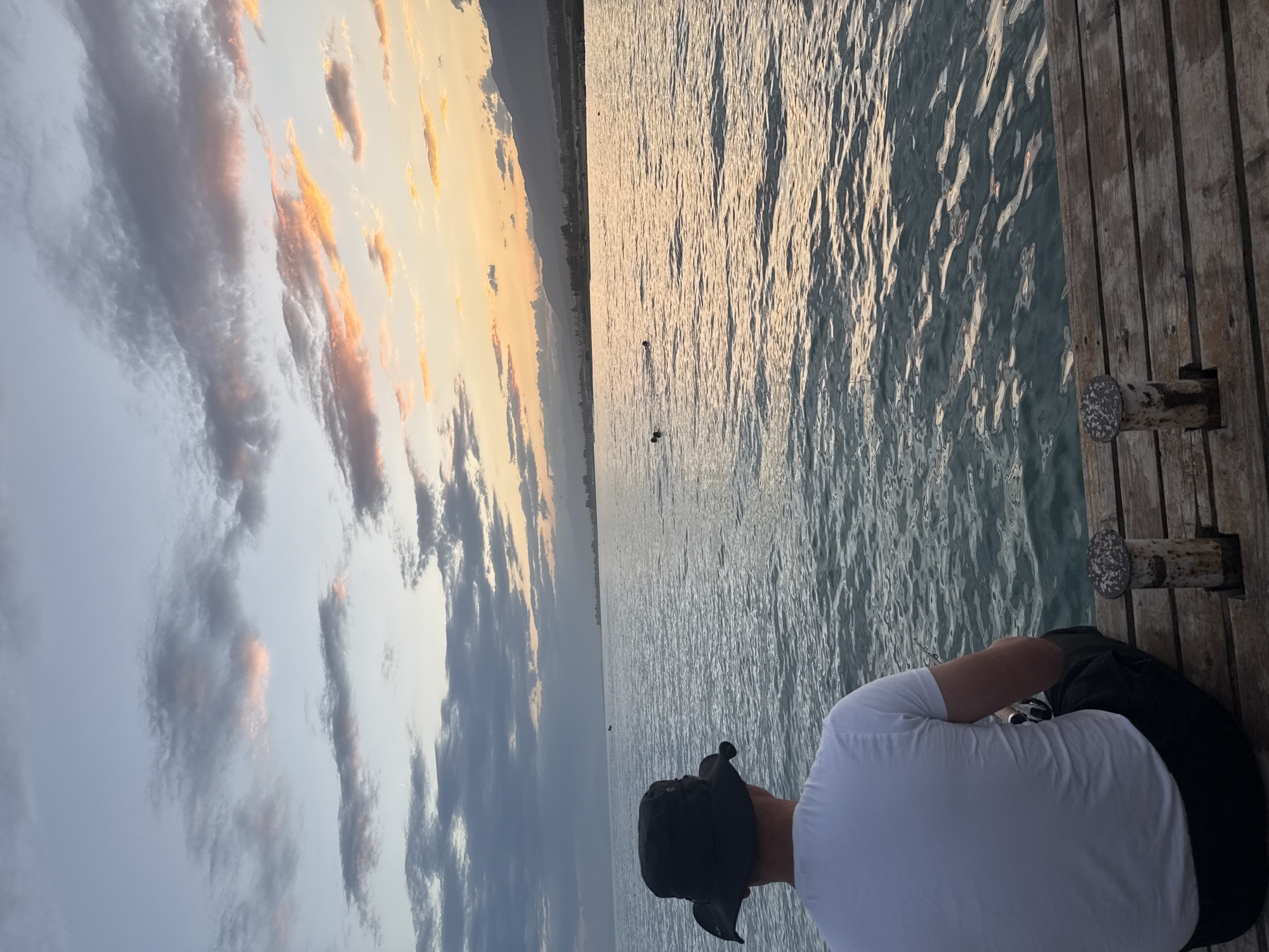 An overhead shot captures a man wearing a white t-shirt and a black bucket hat from behind, sitting on a wooden pier overlooking a calm body of water. The man is positioned in the lower left quadrant of the image. In the distance, across the water, a hazy silhouette of mountains and a town can be seen under a twilight sky filled with scattered clouds tinged with orange and pink hues. Several people are visible swimming or floating in the water further out. Two metal mooring posts are in the foreground on the pier. The scene suggests a serene evening at a lake or sea, potentially near Sary-Oy, Kyrgyzstan, given the mountainous backdrop and the presence of a large body of water.