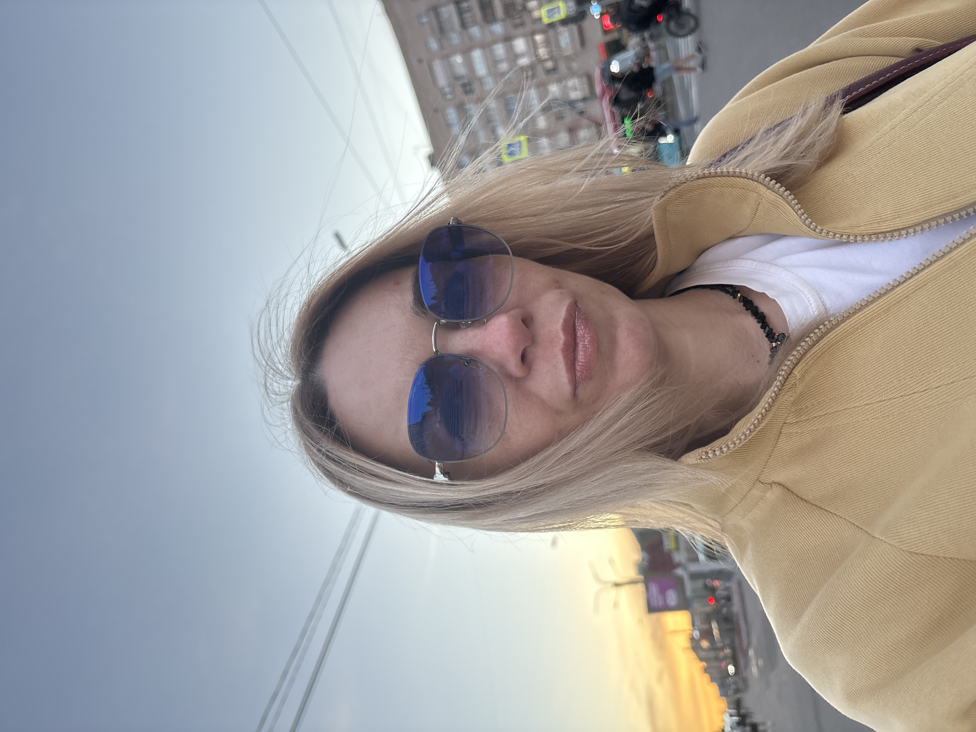 A blonde woman with blue sunglasses and a yellow jacket stands on a street in Sinyavino, Russia. The sun is setting, casting an orange glow on the sky. In the background, buildings and traffic can be seen. The woman is looking at the camera and has a neutral expression.