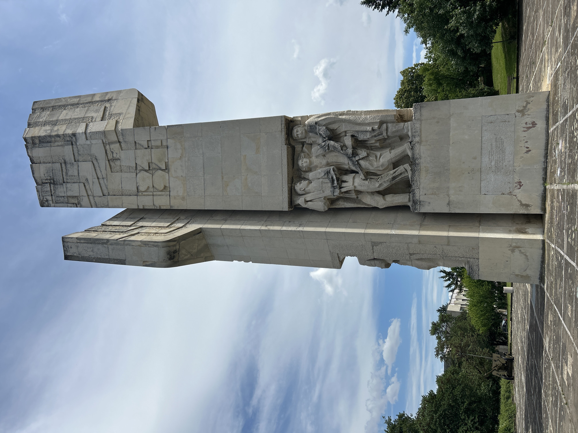 This is a low-angle, full shot of a large, concrete monument with intricate carvings, set in an outdoor plaza. The monument is tall and angular, with geometric patterns etched into its surface. On the front of the monument, there is a bas-relief sculpture of three figures, seemingly marching forward, carrying weapons. The figures are rendered in a stylized, blocky manner. Below the figures, there is a rectangular plaque with an inscription. The plaza is paved with square concrete tiles and bordered by a low wall. To the left of the monument, a vintage artillery piece is visible. Beyond the plaza, there are lush green trees and buildings in the background. The sky above is a mix of blue with scattered white clouds. The lighting suggests it is daytime. The overall scene conveys a sense of history and solemnity.