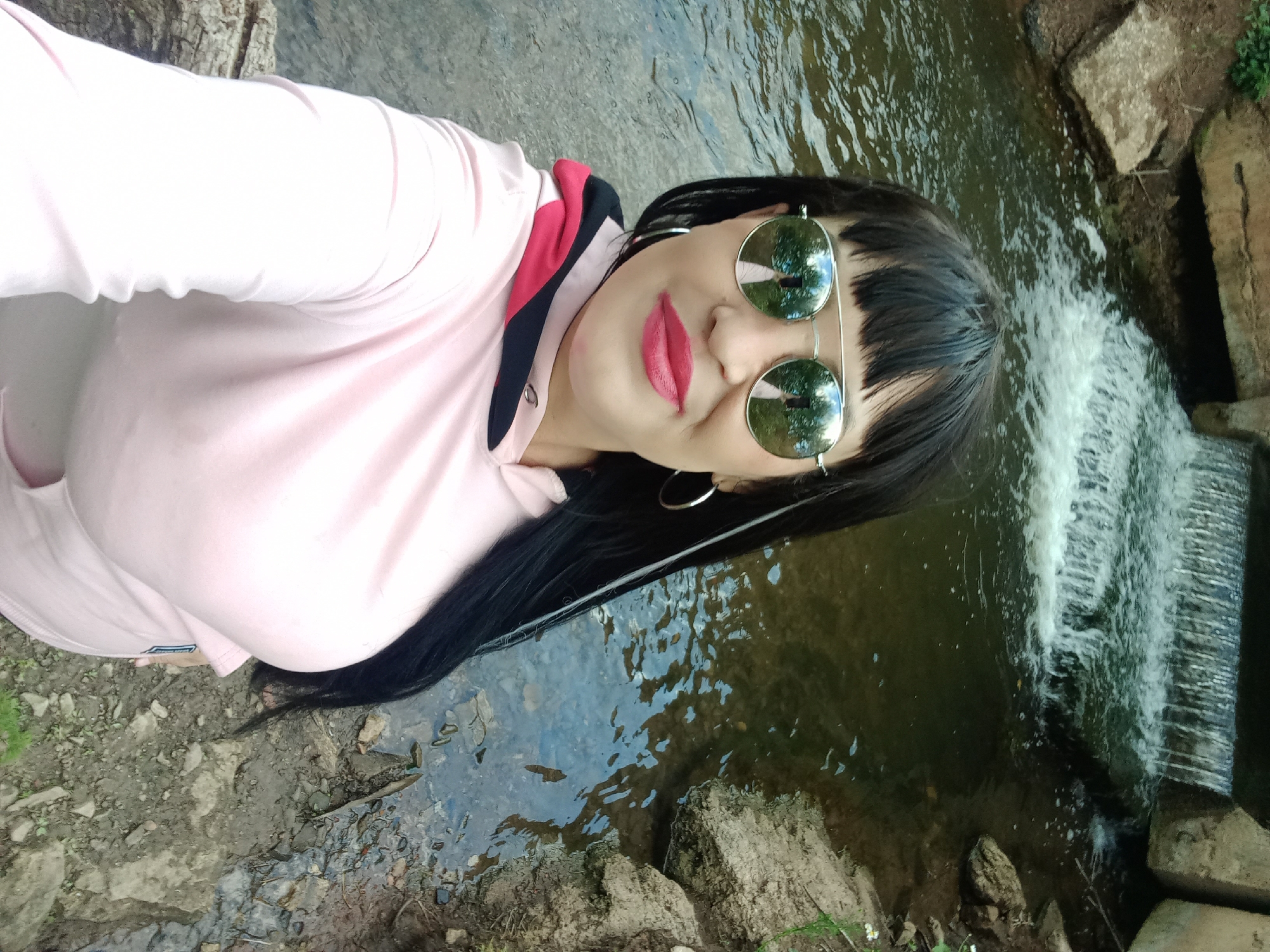 A woman is taking a selfie near a waterfall. The woman has long, black hair, wears mirrored aviator sunglasses and bright pink lipstick. She is wearing a light pink hooded sweatshirt with contrasting black and red trim around the hood. She is standing on a rocky bank next to a body of water that appears to be a stream with a small waterfall in the background. The surrounding environment consists of rocks, dirt, and some greenery. The image is taken from a high angle, looking down at the woman and the scene.