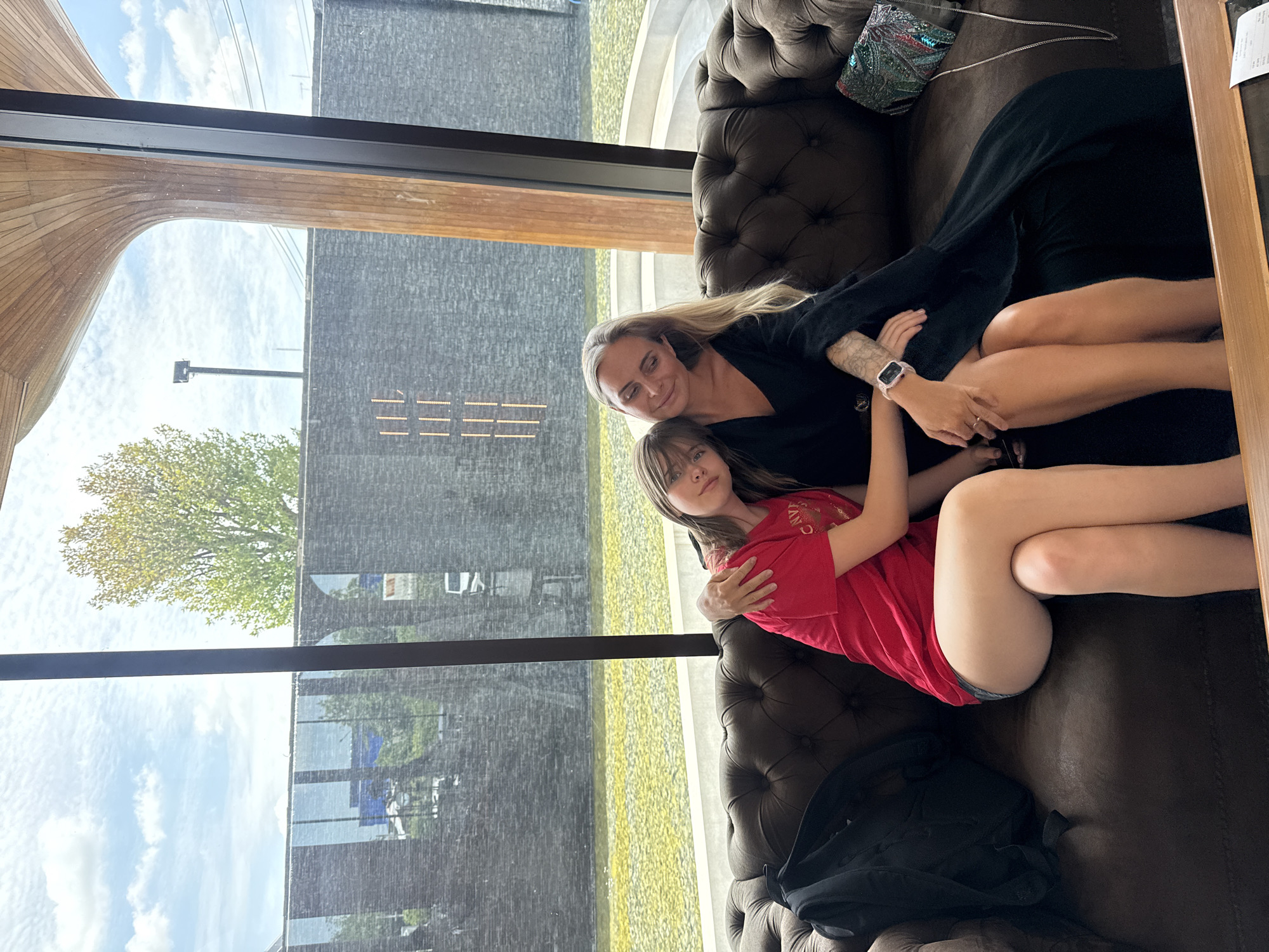 Two women are seated on a dark brown tufted sofa in front of a large window overlooking a bright outdoor area. The woman on the left, appearing younger, wears a red t-shirt and grey shorts, with her left arm around the other woman's waist. The woman on the right, with blonde hair, wears a long black dress and has her right arm around the younger woman's shoulders. Her left hand rests on her lap, and she has a wristwatch on her left wrist. A black backpack is on the sofa to the left of the younger woman, and a beaded purse is on the sofa to the right of the woman in black. The outdoor scene visible through the window features a body of water with a yellow hue and a textured dark stone wall. A large tree with green and yellow leaves is visible on the left side of the frame, and a modern wooden architectural element curves from the top right of the window. The image was taken at the Commune, a co-working space in Bangkok, Thailand.