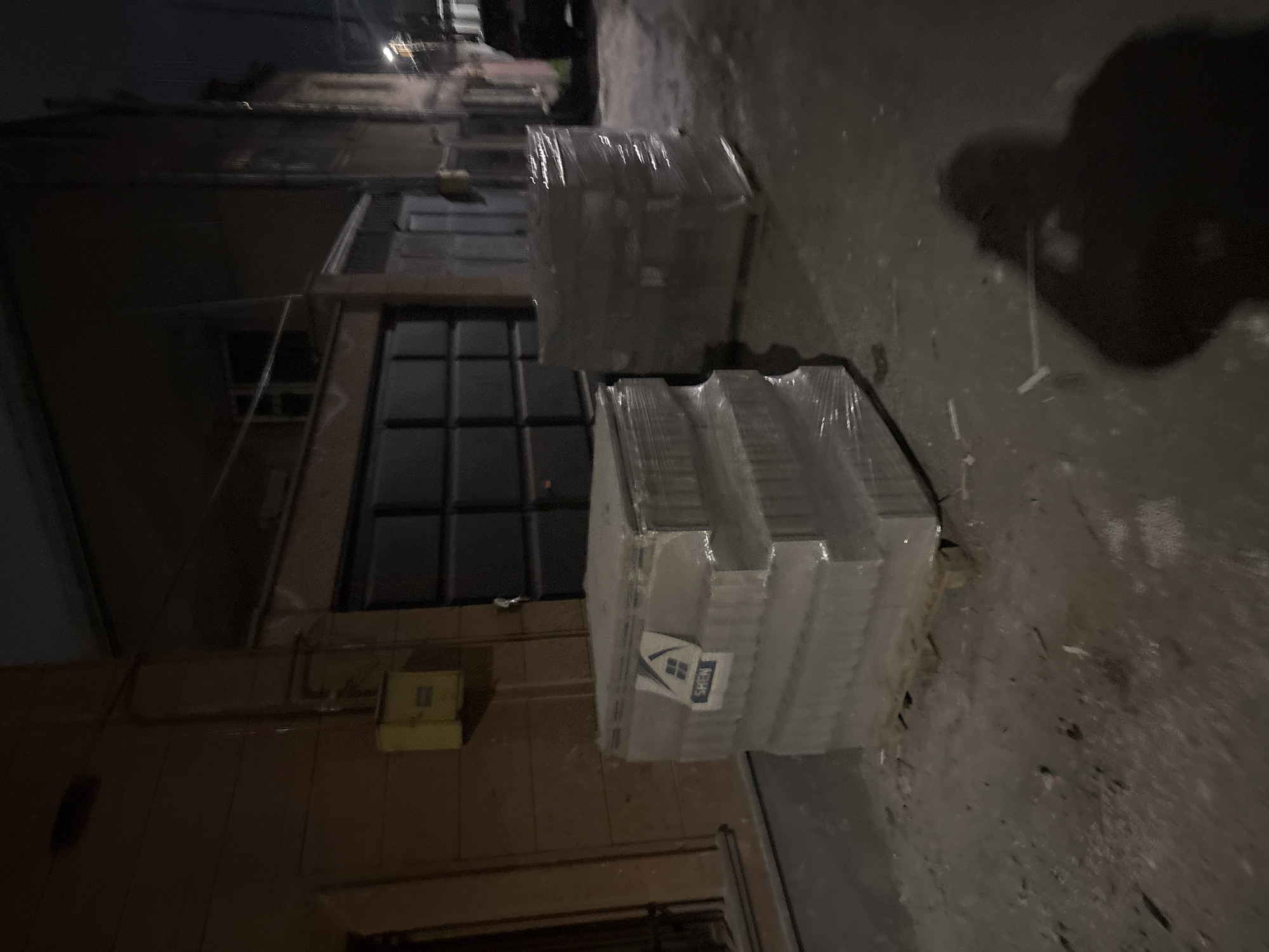 A low-angle photograph depicts a dimly lit alleyway in Yerevan, Armenia. On the left, the exterior wall of a building is visible, featuring a large, dark window divided into multiple panes. A yellow electrical box is mounted on the wall. In the foreground, two stacks of wrapped materials are positioned on the ground. A label on one of the stacks features a logo with a window graphic and the word "SKEN." The ground surface is uneven and appears to be concrete or asphalt. A faint, dark shape resembling a shadow is present in the upper right of the frame. The overall scene suggests a storage area or loading zone.