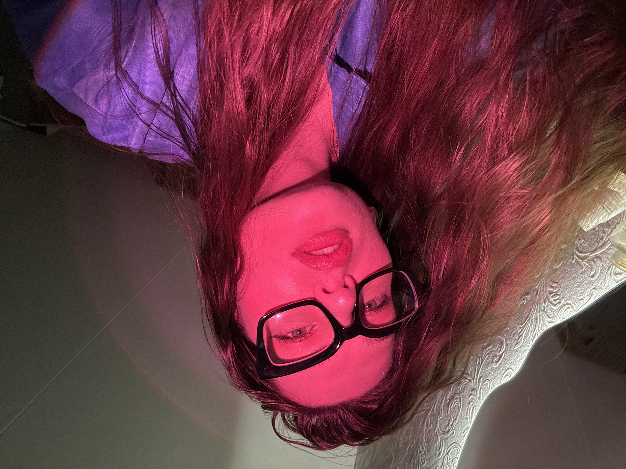 A woman with long, reddish-brown hair and glasses is oriented upside down in the frame. Her face is illuminated with a red light, highlighting her lips and the frames of her glasses. She appears to be wearing a blue or purple top. The background consists of what appears to be a light-colored tiled floor on the left, and an object, possibly a table, covered with a textured white material on the right. There is no information to indicate this photo was taken in Chaikovsky, Russia.