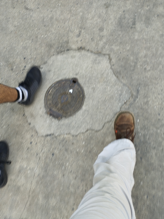 An overhead view captures a section of a concrete or asphalt ground surface in the city of Guacara, Venezuela. Centrally positioned is a circular, dark metal manhole cover, exhibiting signs of wear and a small opening on its upper edge. Two sets of human lower extremities are visible within the frame. To the upper left, a person's lower leg, displaying skin, is present alongside a foot encased in a black athletic shoe with a white sole stripe and a white sock with two black bands. To the lower right, another person's leg, covered by light-colored, wrinkled trousers, terminates in a foot wearing a brown, lace-up shoe with orange-brown accents. Both individuals appear to be standing or moving on the ground, with no direct interaction discernible between them.