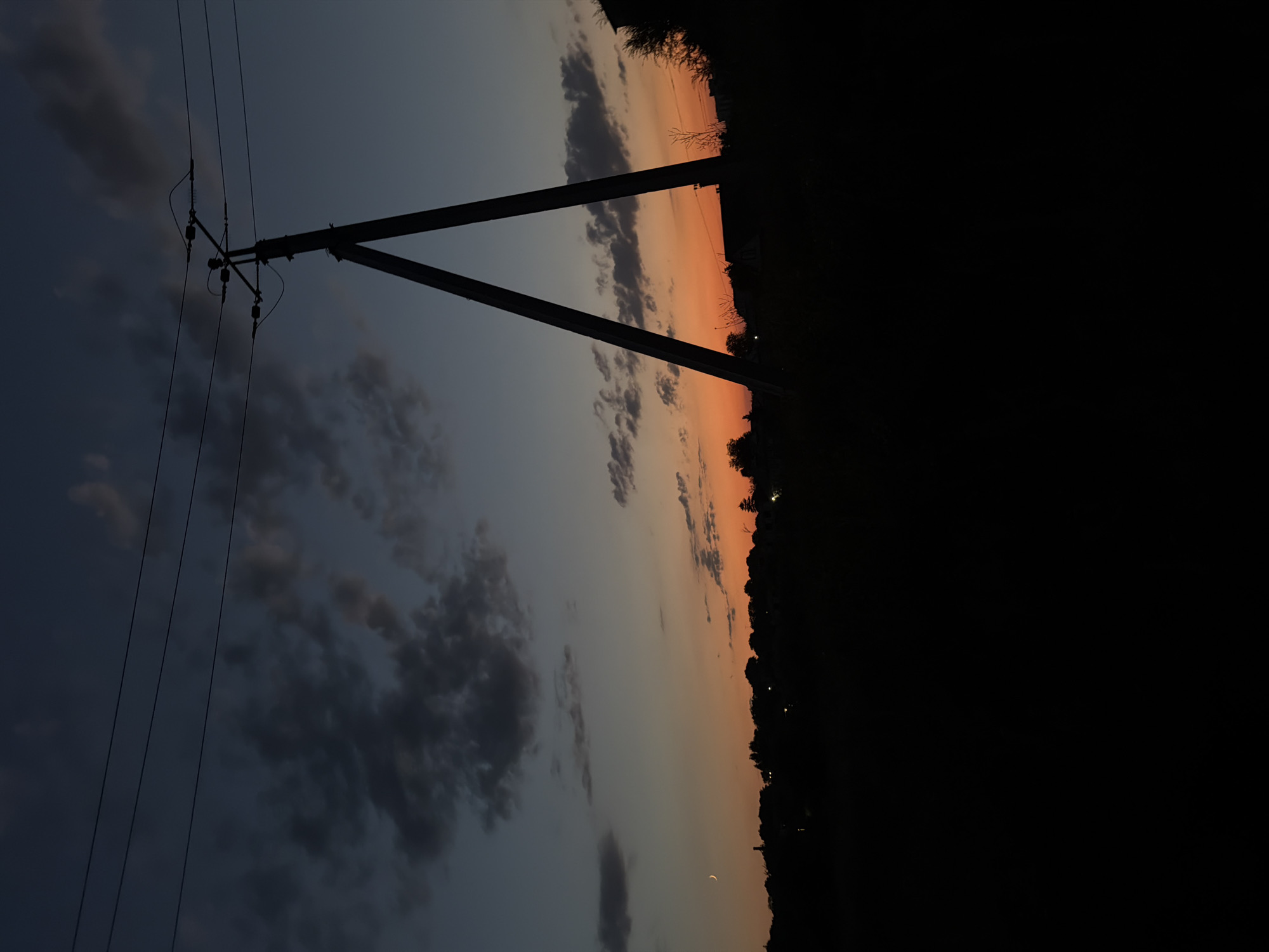 The image captures a silhouetted landscape at dusk in the city of Strășeni, Moldova. In the foreground, dark vegetation and the dark outline of buildings create a stark contrast against the twilight sky. A prominent A-frame utility pole with wires stretches across the upper portion of the frame, silhouetted against the gradient sky. The sky itself transitions from a deep blue at the top to a vibrant orange and yellow near the horizon, indicative of a sunset. Wispy clouds are scattered across the sky, some darker and more defined, while others are lighter and more translucent. A faint crescent moon is visible in the upper left portion of the sky. The overall scene suggests a rural or semi-rural outskirts of the city.