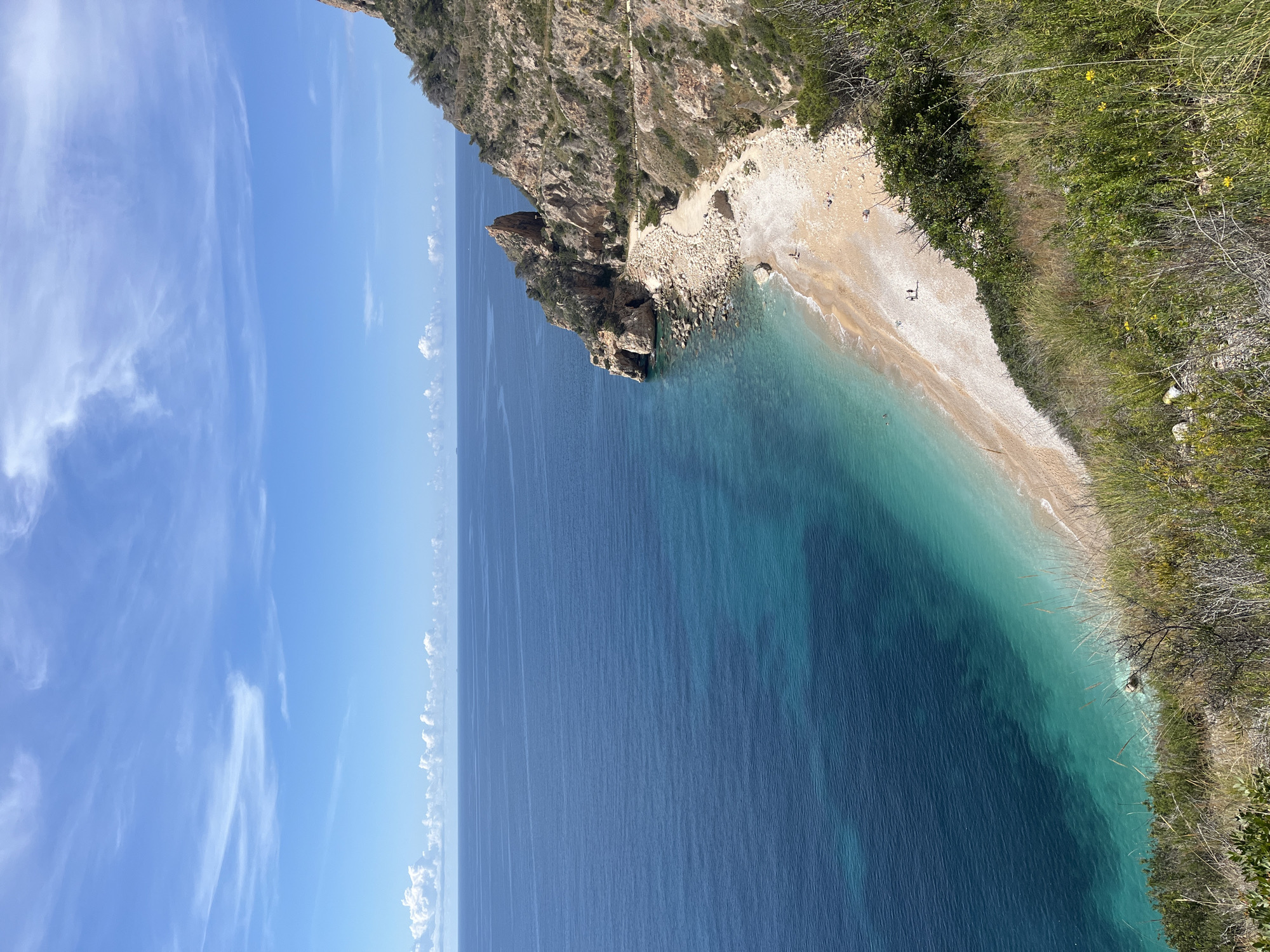 This photograph depicts a coastal scene at Cala del Moraig, a beach in El Poble Nou de Benitatxell / Benitachell, Spain. The composition is divided into three main sections: the sky, the sea, and the coastline. The sky is clear blue with wispy white clouds. The sea is deep blue and turquoise. The coastline consists of a steep, rocky cliff covered with vegetation on the right side. At the foot of the cliff is a pebble beach. Two figures are visible on the beach.
