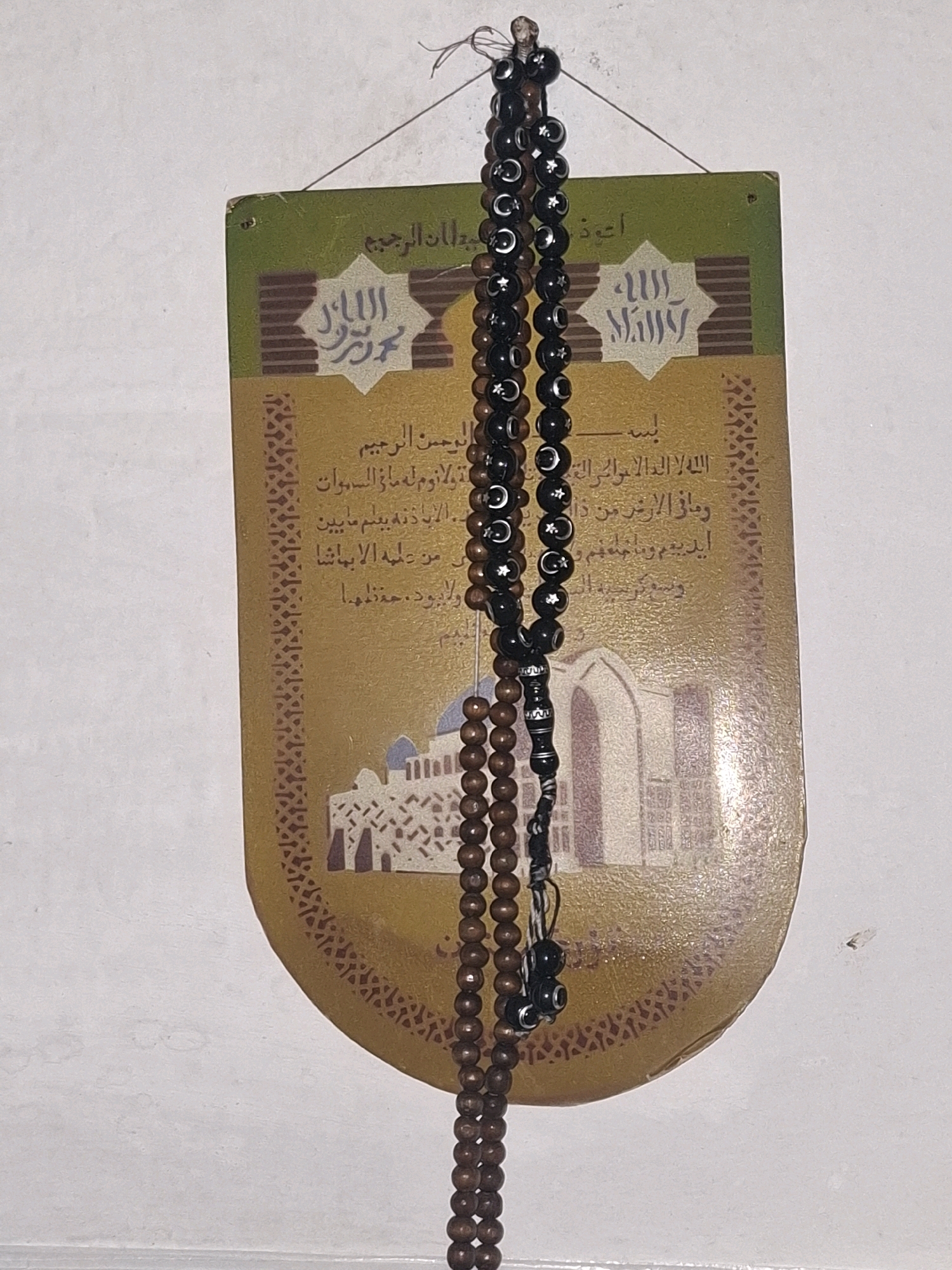 This image shows a close-up of a traditional Islamic wall hanging featuring Arabic calligraphy and prayer beads. The wall hanging is rectangular with a rounded bottom and is predominantly gold and green. The calligraphy appears to be religious text. Two strings of prayer beads are hanging in front of the wall hanging. One string is made of dark, possibly black, beads with crescent moon and star designs. The other string is made of lighter brown, wooden beads. The background is a plain, white wall. The image is taken from a slightly elevated angle, looking down at the objects. There are no people or animals present in the image, and no specific identifiable landmarks are visible to determine a location in Almaty, Kazakhstan.