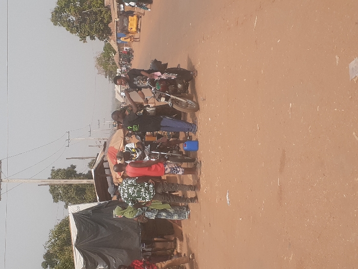 The image displays an unpaved street scene in Gwadanye, Nigeria, under a hazy, light-gray sky. The foreground consists of reddish-brown dirt. Approximately ten individuals are visible, predominantly adult males. On the left, a person in a light green head covering and patterned attire stands near a makeshift market stall covered with a black tarp. Adjacent to this, multiple individuals are gathered, including one in a camouflage-patterned shirt and another in a red tank top, positioned near a motorcycle.
In the central area, two men are actively interacting between two motorcycles. One man, wearing a black T-shirt with a green logo on the chest and blue trousers, gestures with his right hand towards another man in a black T-shirt, who stands next to a dark-colored motorcycle. A blue plastic jerry can rests on the ground at their feet. The background reveals a dirt road extending into the distance, flanked by low-rise structures and sparse vegetation. Multiple wooden utility poles with overhead power lines are prominent. A yellow motorized rickshaw is visible further down the road on the right side.