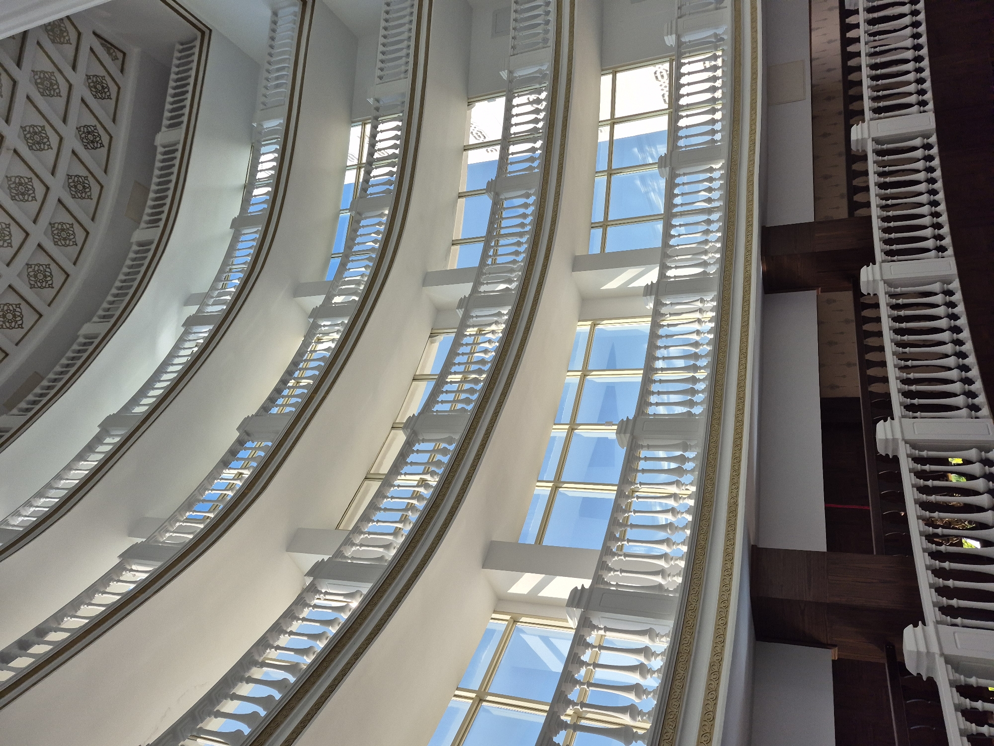 This image depicts an interior architectural view, most likely of the Alanya Culture Center in Alanya, Turkey. The scene is oriented upwards, focusing on the curved white walls adorned with balustrades and windows. The windows reveal a clear blue sky. The architectural style is ornate, featuring decorative gold trim along the walls. On the right side, a dark wooden structure is visible, also trimmed with white balustrades. The lighting is bright, illuminating the predominantly white and light surfaces.