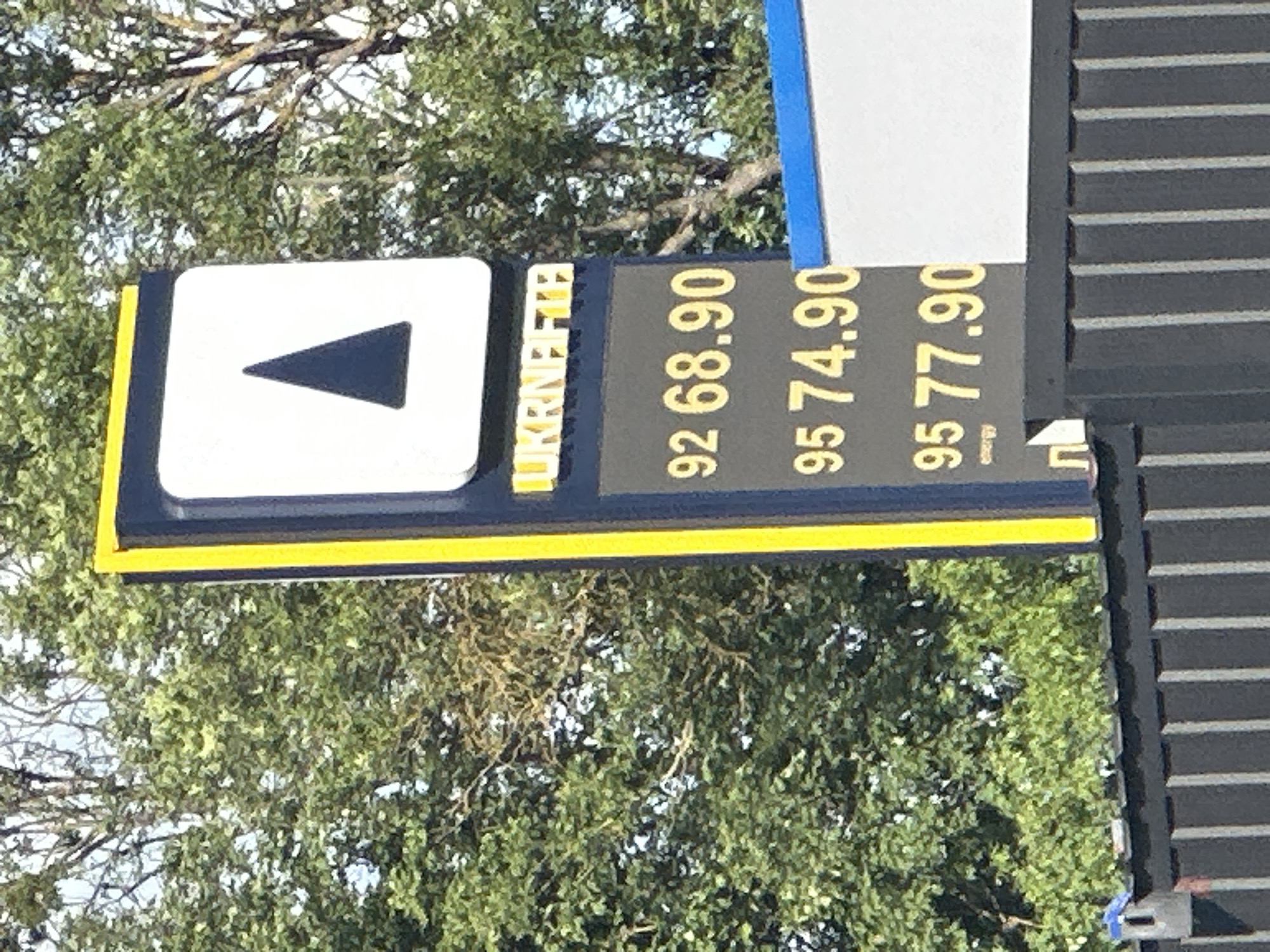 The image shows a gas station sign in Shatava, Ukraine. The sign has a white square with a black triangle in the center, and the words "UKRNAFTA" in yellow letters below it. The sign also displays fuel prices: 92 octane at 68.90, 95 octane at 74.90, and another 95 octane at 77.90. The sign is tall and slender, with a yellow border. The background consists of green trees and a blue sky. A dark fence with vertical slats runs along the bottom of the image.