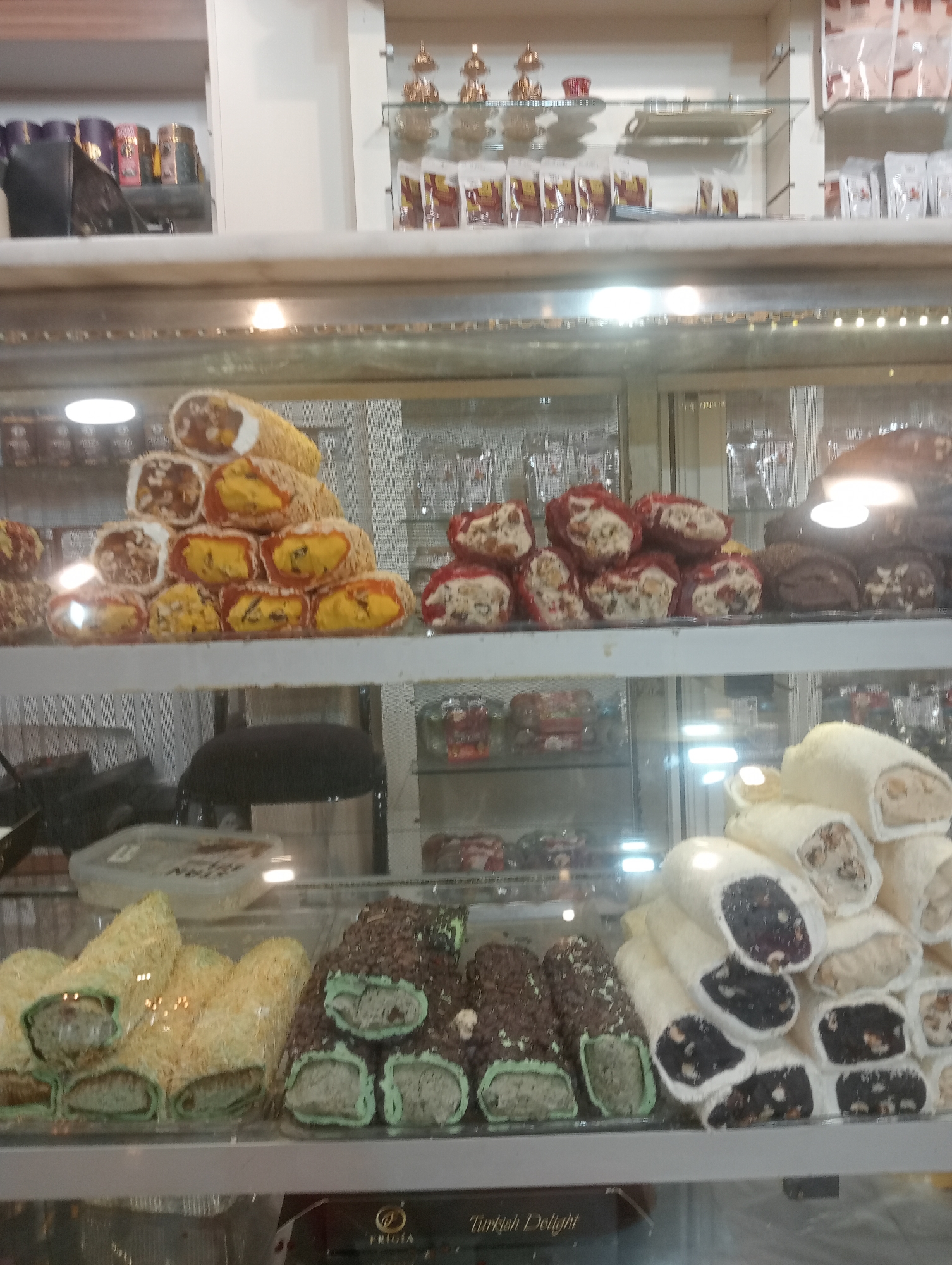The image shows a display case filled with Turkish Delight, a confection. The display case is in a shop. The Turkish Delight is of various types, some rolled and covered in nuts or coconut, others cut into cubes and coated in powdered sugar. The shop appears to be in the city of Korkuteli, Turkey, as indicated by the label "Turkish Delight" on a box in the lower part of the image. There are no people visible in the image. The lighting in the display case is bright and reflects off the glass. The overall impression is of a well-stocked sweet shop.