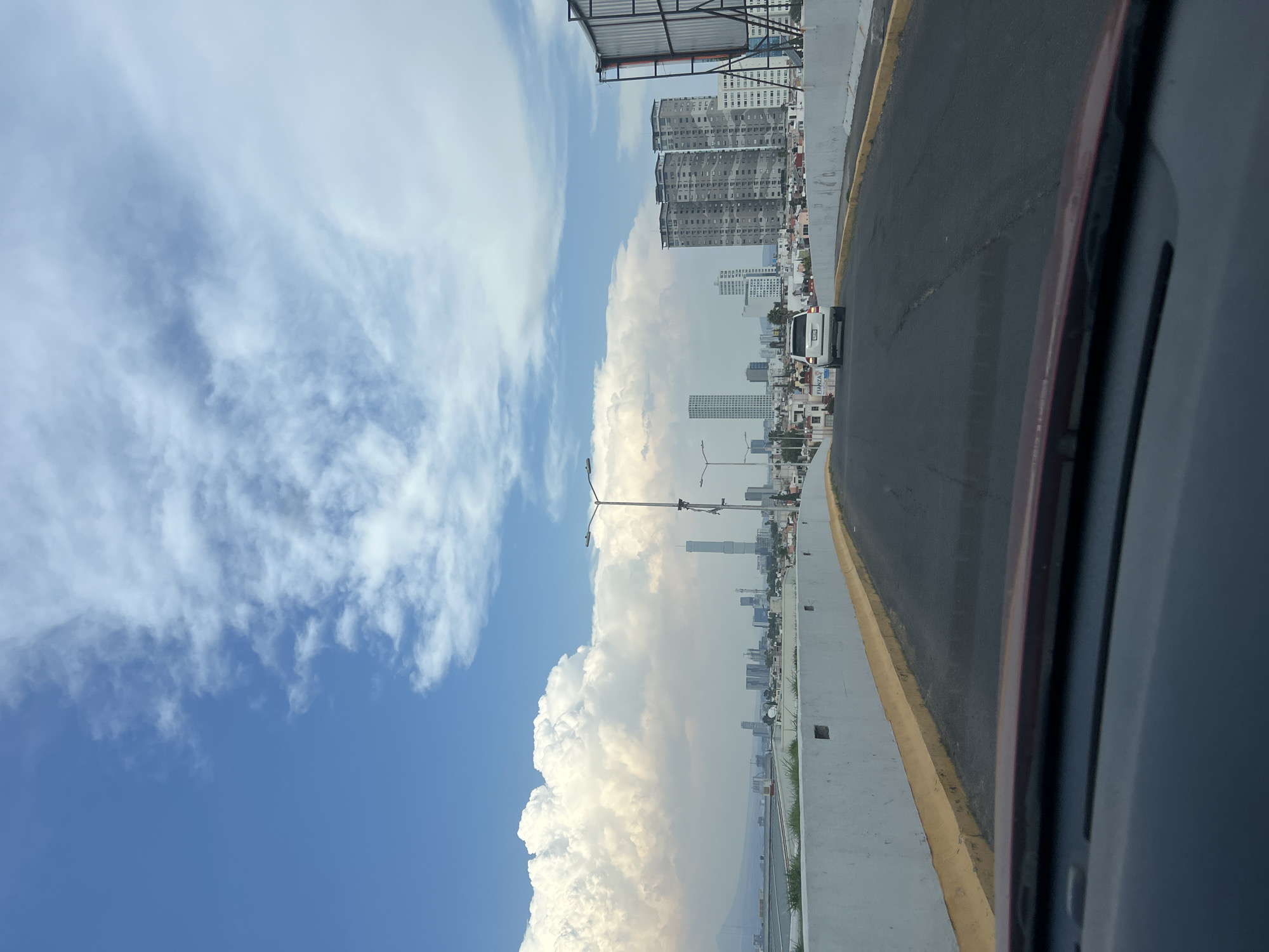 The image depicts a view from a vehicle in San Bernardino Tlaxcalancingo, Mexico. To the left, a blue sky is filled with white clouds. In the distance, a cityscape with tall buildings and smaller structures stretches toward the horizon. A highway or roadway occupies the right side of the frame. The edge of the car window is visible in the right foreground. The buildings and city skyline are consistent with a typical urban environment in Mexico. The light pole in front of the buildings indicates that this area has infrastructure for street lighting. The clouds suggest it's a sunny day with potential for changing weather conditions.