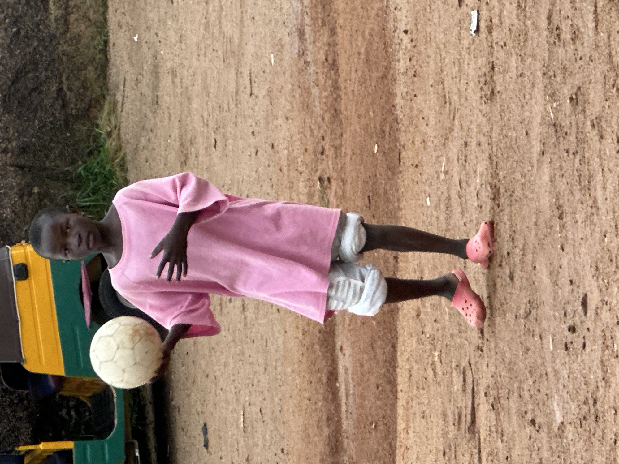 A young Black child stands in an outdoor, dirt-covered area, holding a white soccer ball with their left hand. The child is wearing a bright pink, oversized t-shirt and white, puffy shorts. They are also wearing pink Croc-style shoes. In the background, a yellow, green, and brown motorized tricycle is visible. The ground is composed of reddish-brown dirt with small stones and debris scattered across it. The lighting suggests an overcast day. The scene appears to be in a casual, informal setting, possibly a residential or play area in Jos, Nigeria.