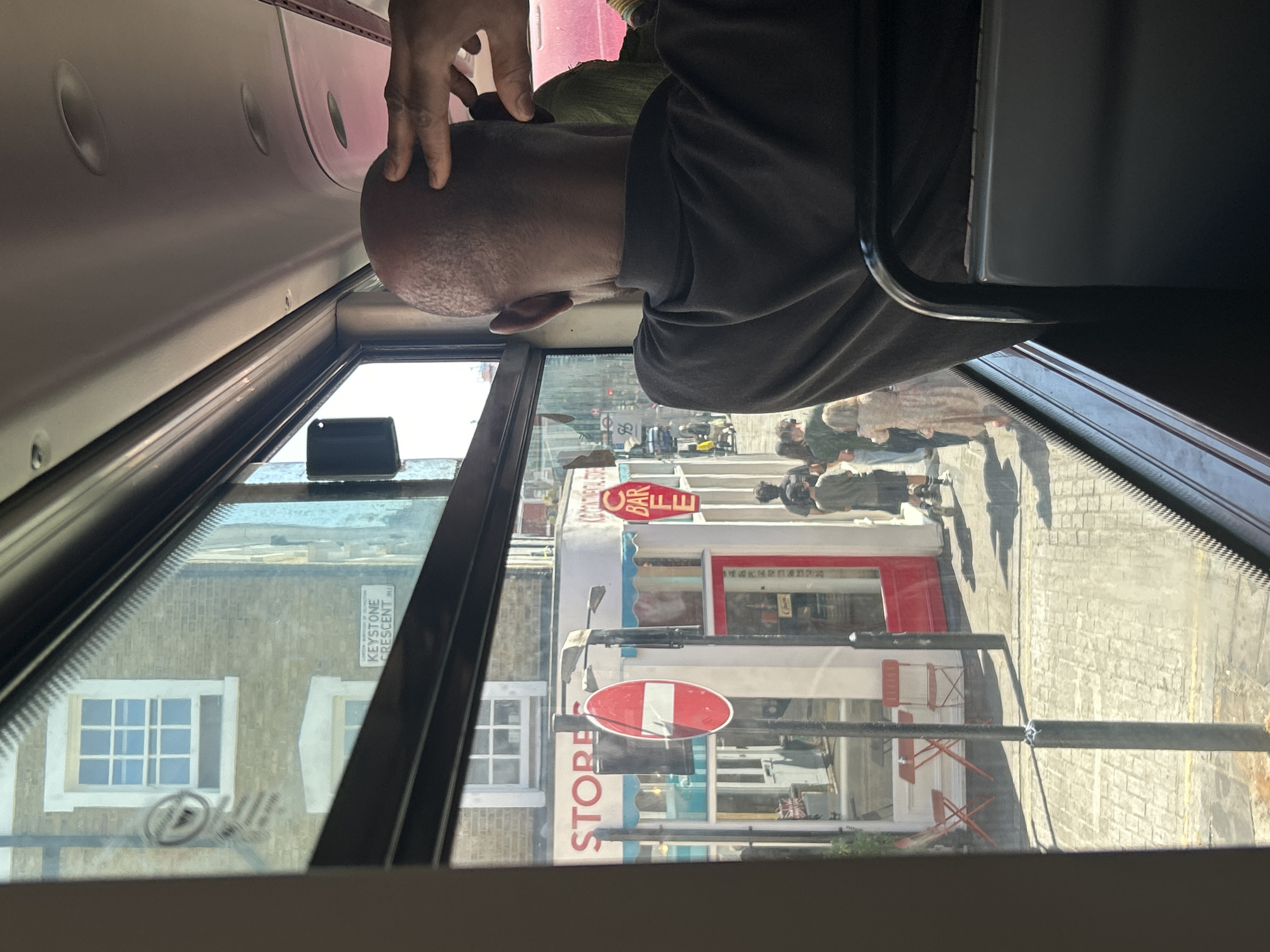 The image, taken from the interior of a bus, offers a view through the window of a street scene in London, United Kingdom. In the foreground, the back of a bald Black man's head is visible, with his hand touching his forehead. Outside the bus, several shops line the street. One shop has a sign that reads "STORES," with red awnings above. Adjacent to it, another establishment features a "CAFE BAR" sign. Pedestrians are walking on the sidewalk in front of the shops. A red and white circular "no entry" sign is prominently displayed on a pole. The street is paved with cobblestones. In the background, a street sign reads "KEYSTONE CRESCENT." The architecture appears to be traditional British brickwork. The lighting suggests it is daytime with bright sunlight.
