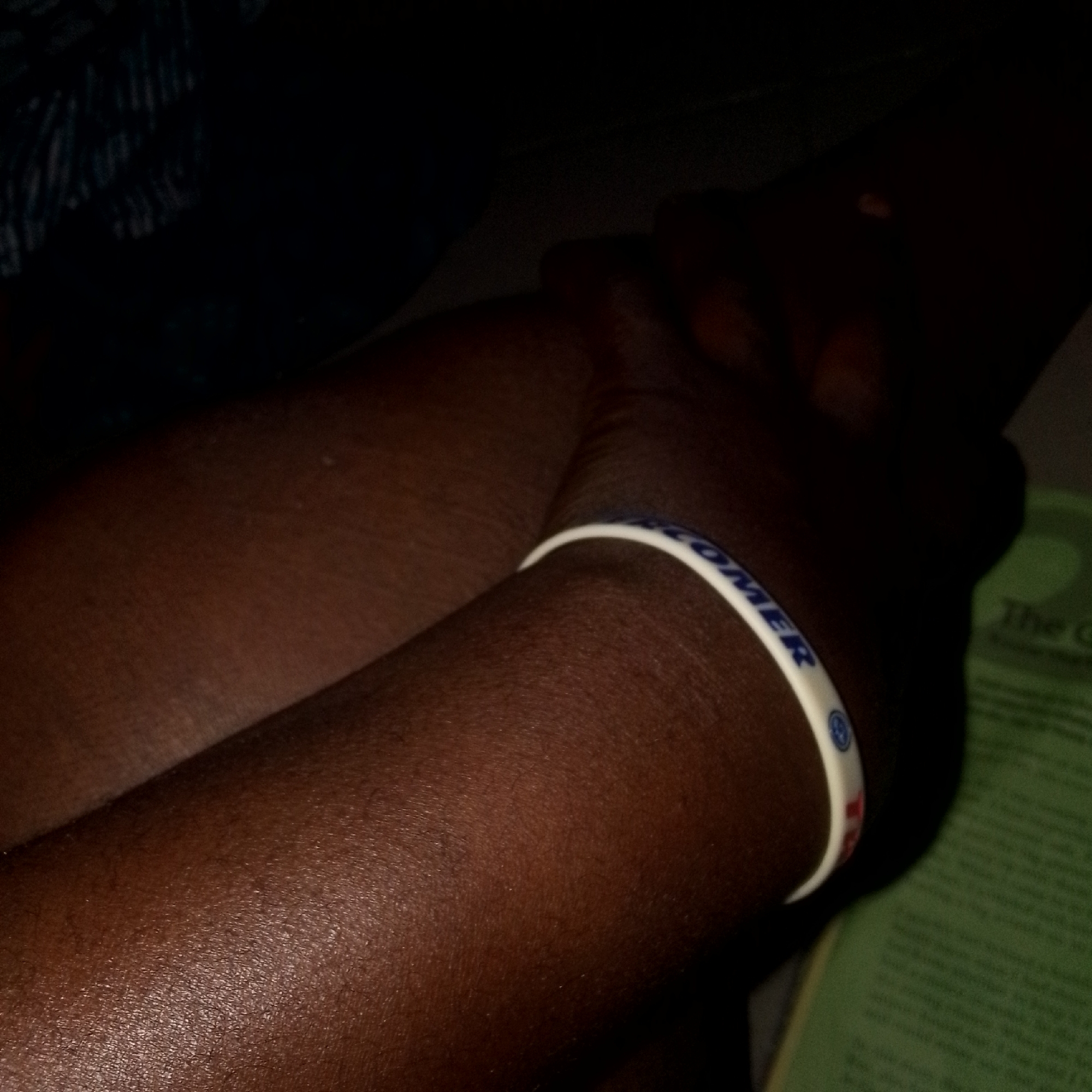 A close-up, indoor shot focuses on the lower arms and wrists of a person with dark skin. The person is wearing a white wristband with blue lettering that reads "CUSTOMER". The wristband also features a blue logo. The person's arms are resting against a green textured surface, possibly a book or magazine, with some text visible. The lighting in the image is dim, casting shadows and highlighting the texture of the skin.