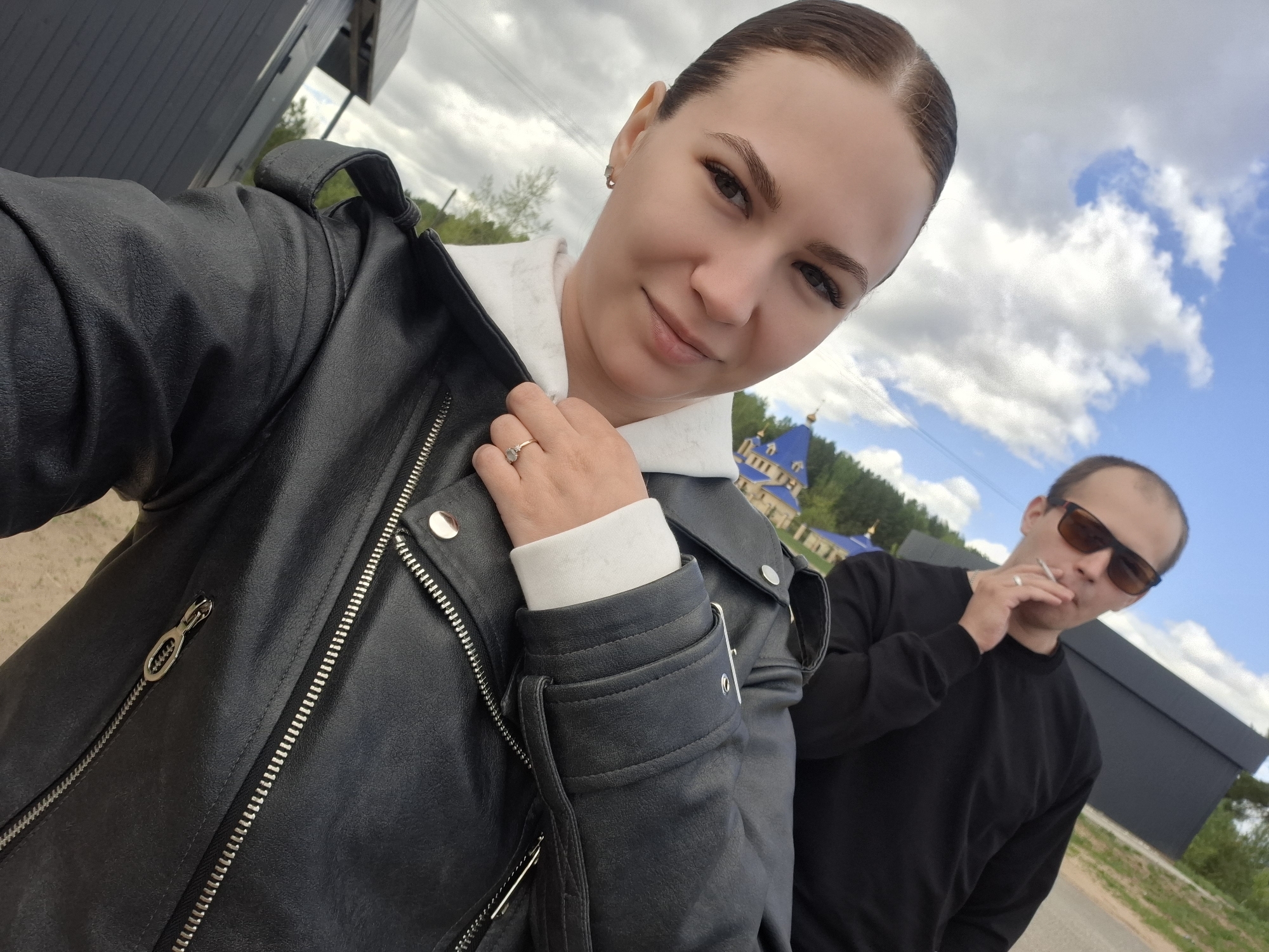 A woman wearing a black leather jacket over a white hoodie is taking a selfie outdoors. She is fair-skinned with her hair pulled back. A ring is visible on her left ring finger. Behind her is a man in sunglasses, dressed in black, holding a cigarette in his mouth. The background shows a partially cloudy blue sky, trees, and a building with blue and gold domes, indicative of a church or religious structure. Other structures resembling modern buildings are also in the background. The location is in Chaikovsky, Russia.