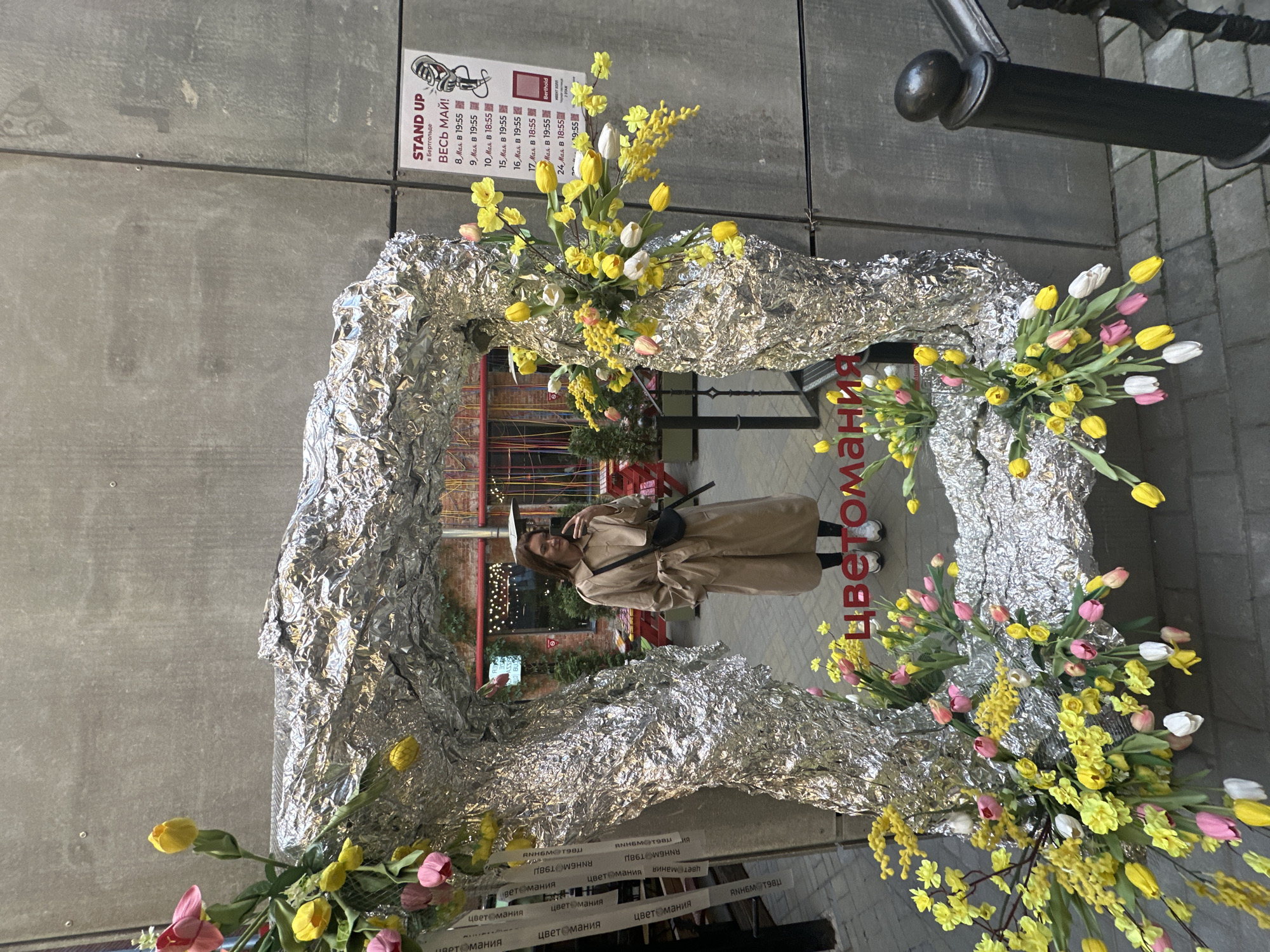 Here's a detailed description of the image:
**General Overview:**
The image shows a person standing within a decorated archway, likely an art installation or a temporary structure for events or promotion. The scene appears to be outdoors, possibly a street or public space.
**People:**
There's one person visible. They are wearing a tan trench coat, black shoulder bag, and white sneakers. They are facing the camera, appearing to be posing or taking a picture.
**Subjects and Objects:**
* **Archway:** The archway is the most prominent feature. It's covered in wrinkled silver foil, giving it a reflective, textured appearance.
* **Flowers:** The archway is decorated with bunches of flowers. There are tulips (yellow, pink, and white) mixed with yellow mimosa.
* **Signage:** On the wall behind the archway, there's a sign in Russian that says "STAND UP".
* **Environment:** The background shows a building with grey panels.
* **Text:** There is text below the archway that spells out "цветомания" multiple times.
**Actions and Interactions:**
* The person is posing or taking a picture within the archway.
**Location:**
While the image alone does not confirm the exact location, one can infer that it is most likely in the city of Saint Petersburg, Russia due to the Russian text.