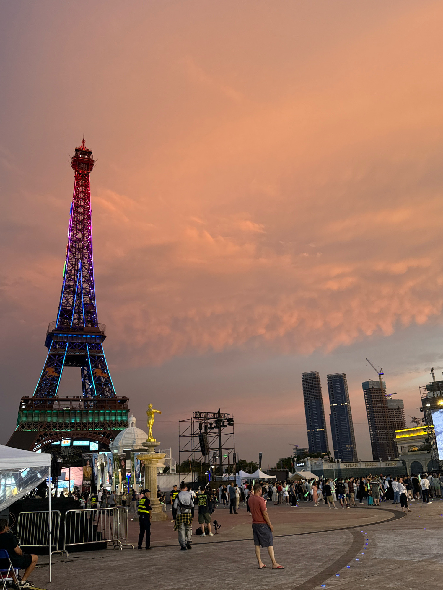 The image depicts an outdoor plaza in Nanshan District, China, at dusk. A replica of the Eiffel Tower, illuminated with colorful lights, stands prominently on the left side of the frame. The sky is filled with orange and pink clouds, suggesting a sunset. In the background, several modern skyscrapers and construction cranes are visible, indicating an urban development area.
The plaza is populated with a crowd of people, some holding glowing sticks, indicating a possible event or festival. Several individuals are dressed in casual attire. Security personnel, identifiable by their vests, are also present, monitoring the crowd. A white tent structure and temporary fencing are set up on the left, possibly for event access or a stage. In the center of the plaza, a golden statue on a fountain is visible. The ground is paved with stone, with some illuminated blue lights embedded in the surface. The overall scene suggests a lively public gathering at an entertainment or cultural venue.