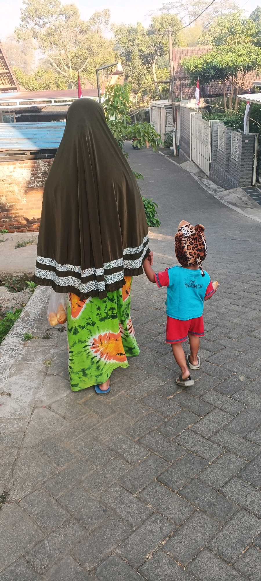 In Penanggungan, Indonesia, a woman wearing a dark brown hijab and a brightly colored tie-dye dress walks on a paved sidewalk, holding a plastic bag filled with bread in her left hand. She is accompanied by a young child dressed in a blue t-shirt and red shorts, wearing a leopard-print hat. The child is walking slightly ahead of the woman, holding a piece of bread in their right hand. The background reveals a residential street with houses, fences, and lush green trees, bathed in warm, natural light. The overall scene depicts a common, everyday moment of a woman and child strolling outdoors in a neighborhood setting.